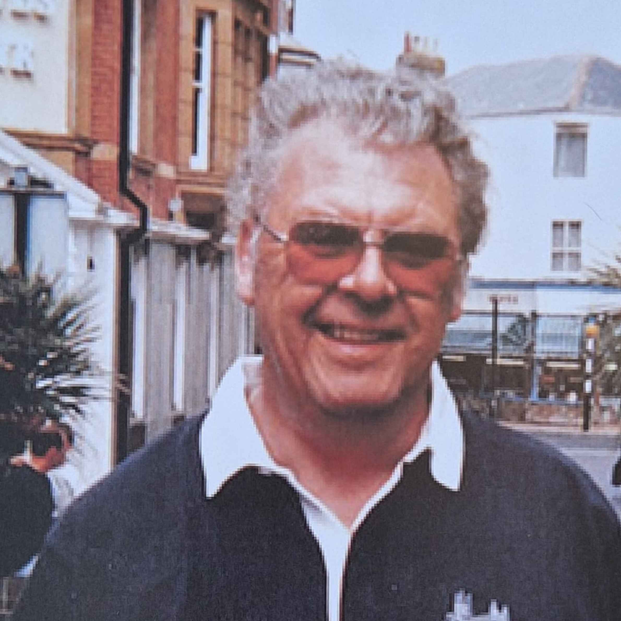 Man with grey hair, wearing sunglasses and smiling, navy polo shirt with white collar. 