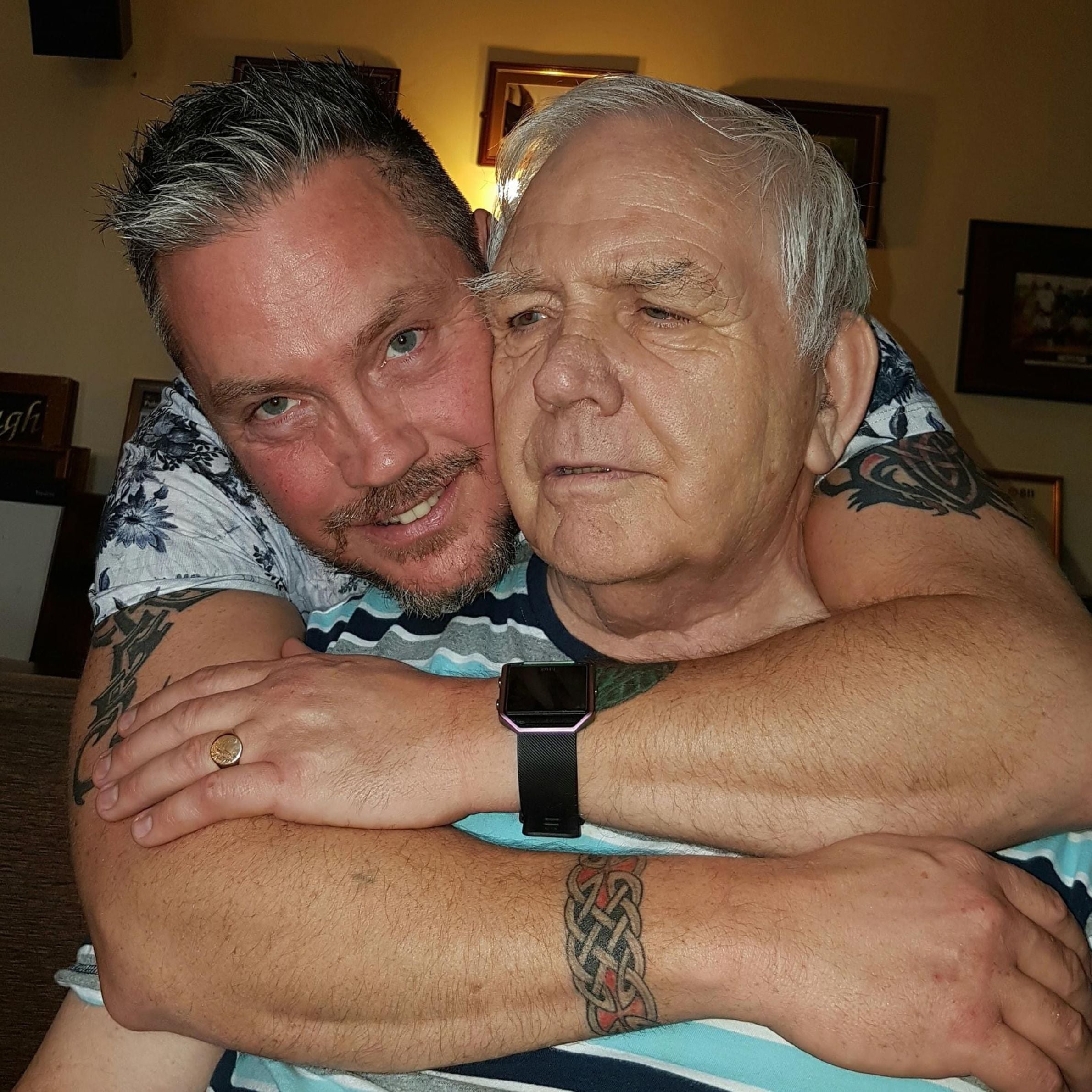a man with short black and grey hair wearing a light grey patterned top and a black watch behind an older man wearing a light blue shirt with his arms over his shoulders.