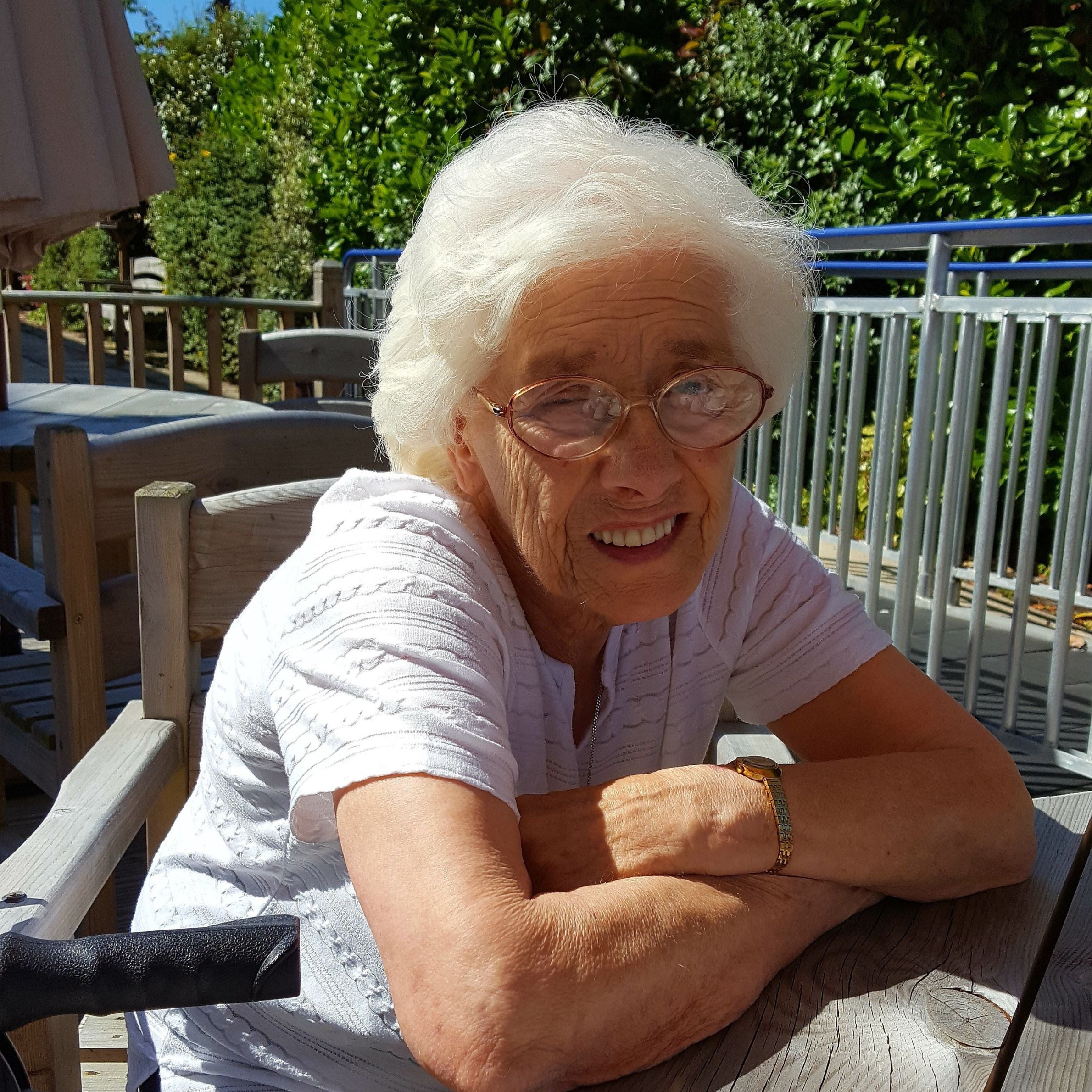 Older lady sat at a table outside in the sunshine. She is smiling and is wearing glasses and a white tshirt. There is a metal fence and trees in the background.