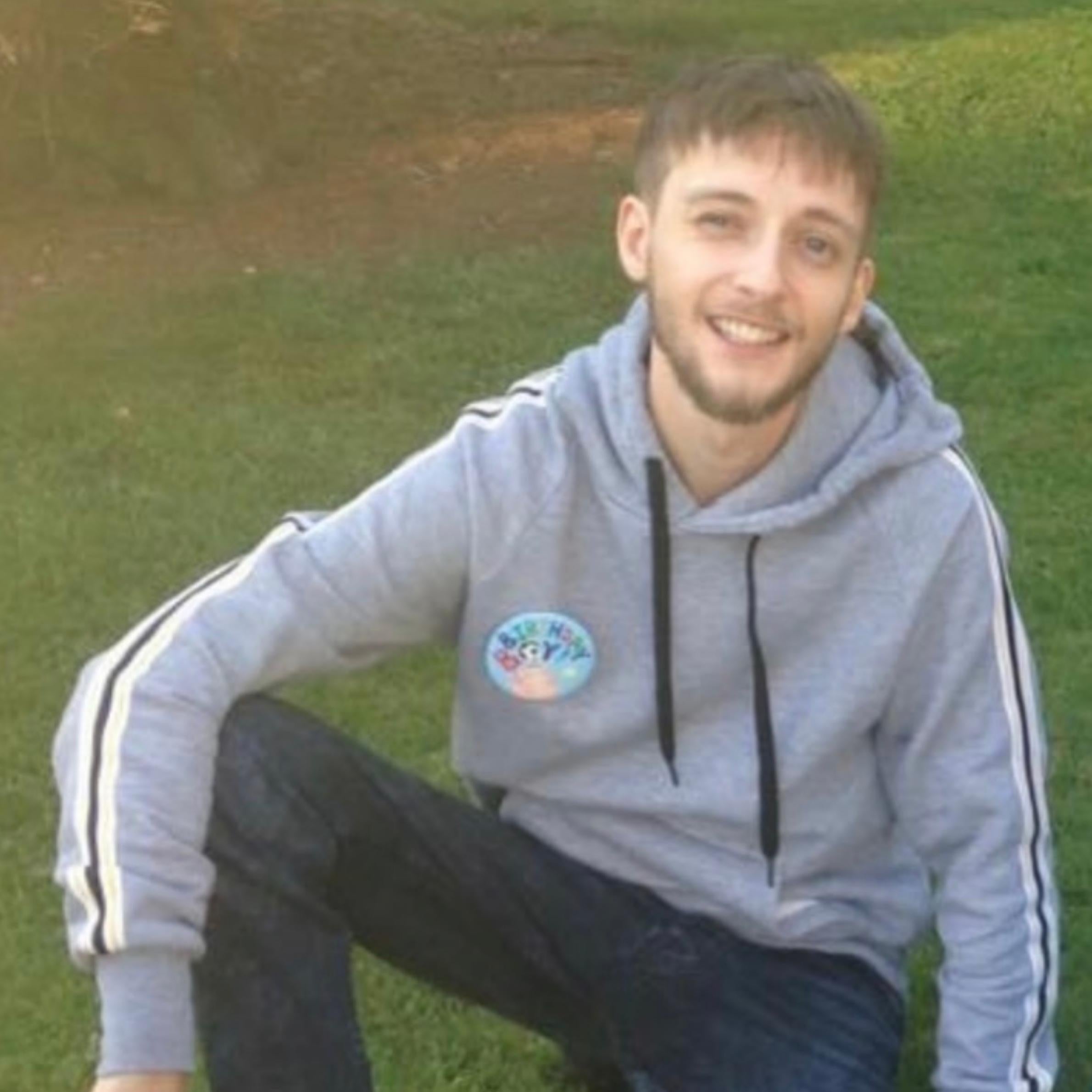 Man wearing grey hoodie and birthday badge. He has dark trousers on. He has brown hair and a brown beard, he is sat on grass. 