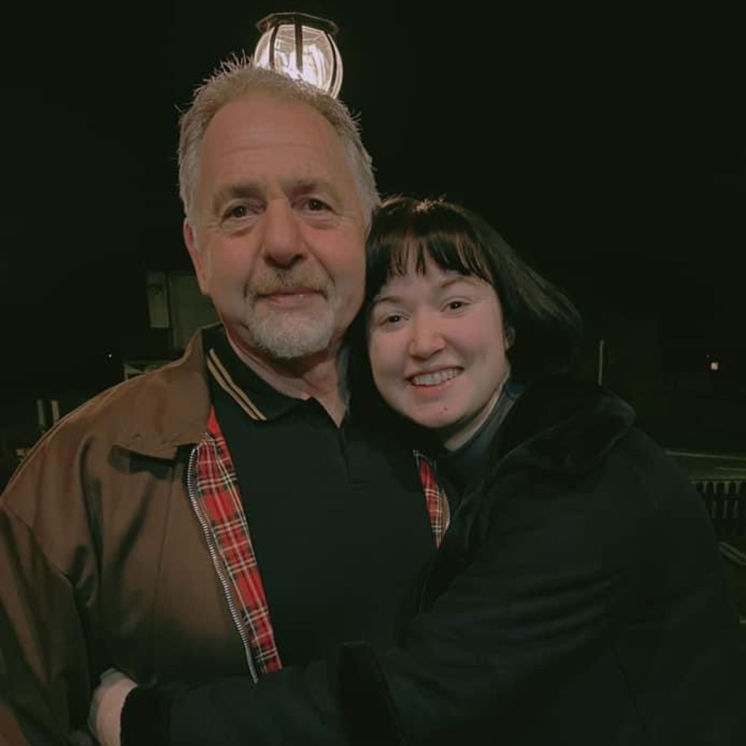 a woman on the right side with black hair wearing a black coat smiling hugging a man on the left wearing a brown and red coat, he has light grey hair and beard. there is a light above their heads in the background
