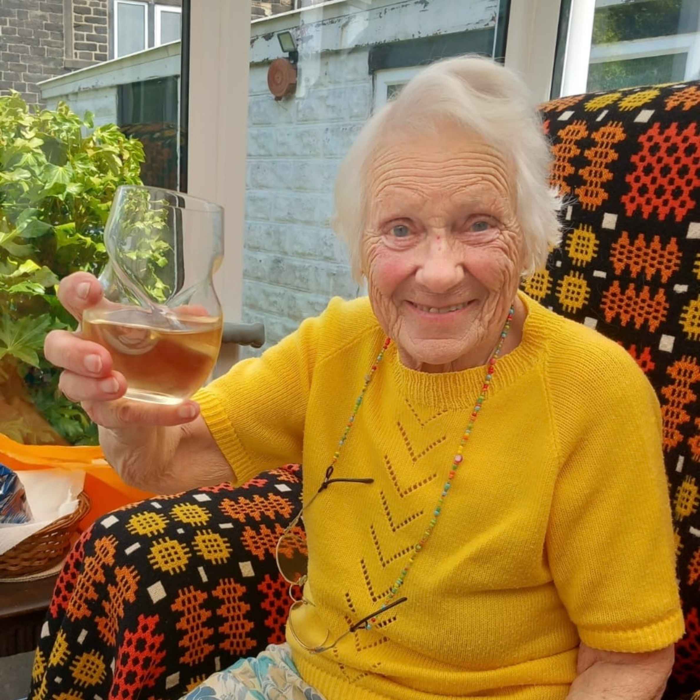 lady smiling holding a glass. She is wearing a yellow knitted short-sleeve jumper. She has short white hair and is sat on a chair with a patterned throw on it. 