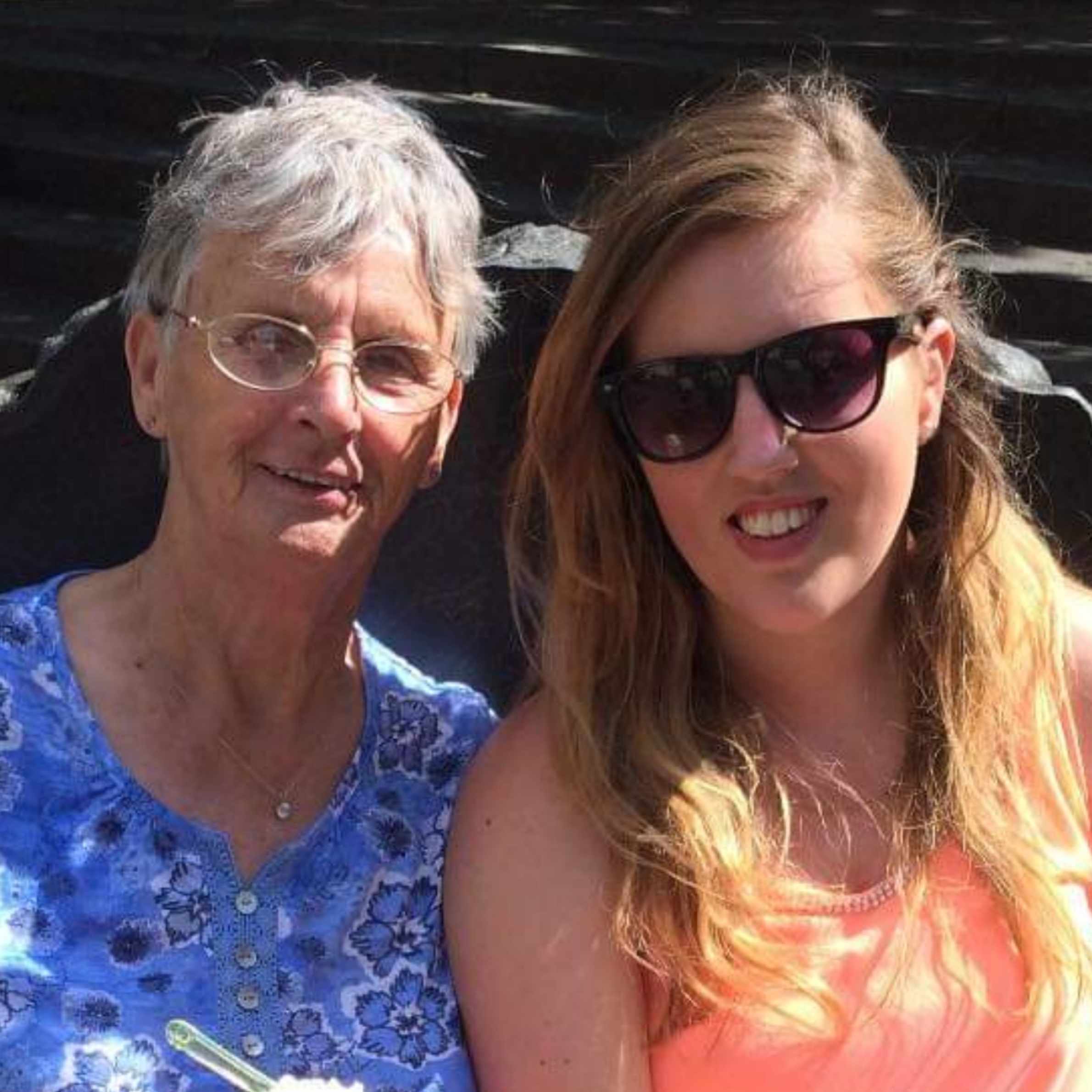 Lady to the left is wearing blue floral blouse, she has short grey hair and is wearing glasses. Lady on the right is wearing a coral coloured vest top and sunglasses. She has long brown hair. 