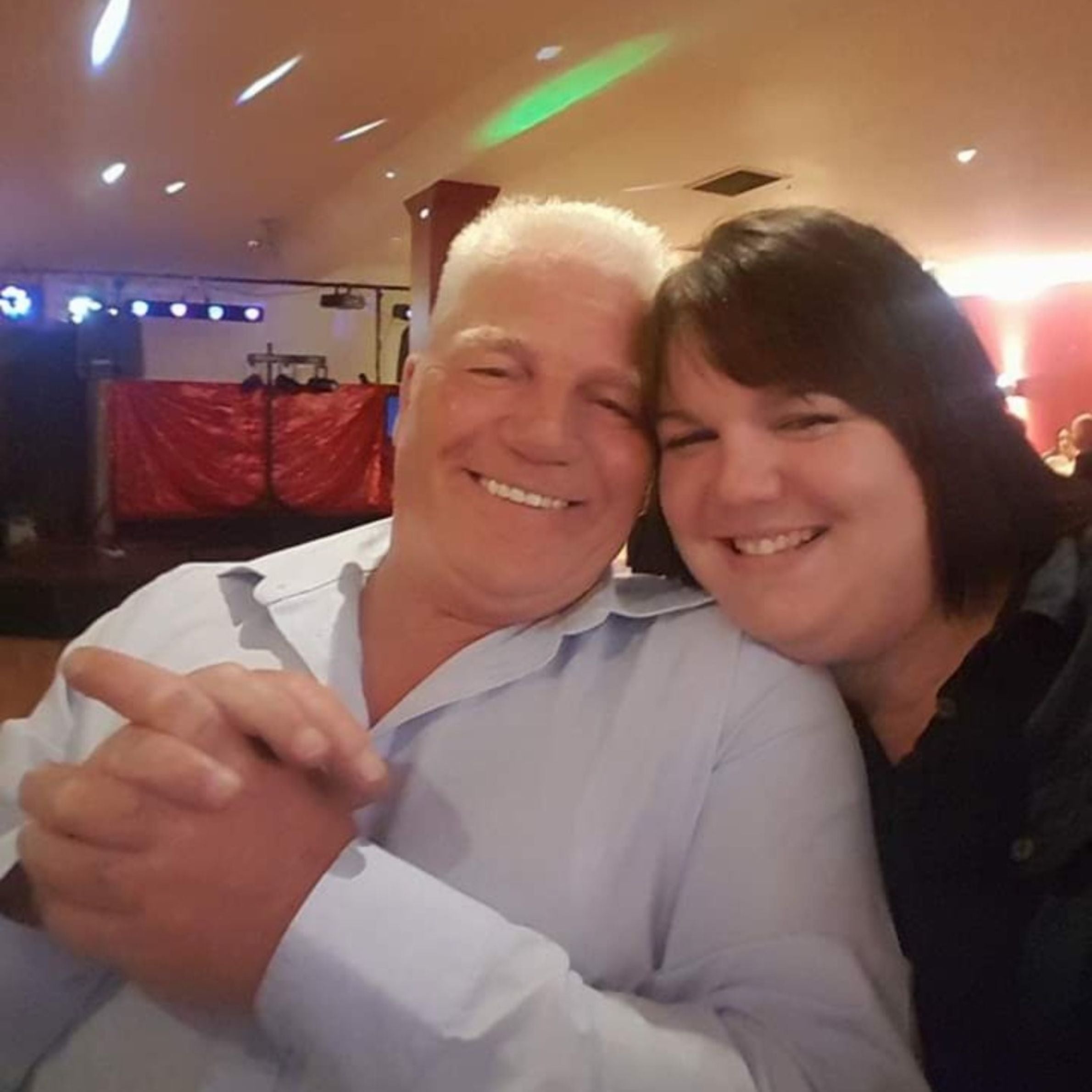 A man in a blue shirt with white hair next to a woman in a black shirt with dark hair smiling. There is a red curtain in the background and disco lights
