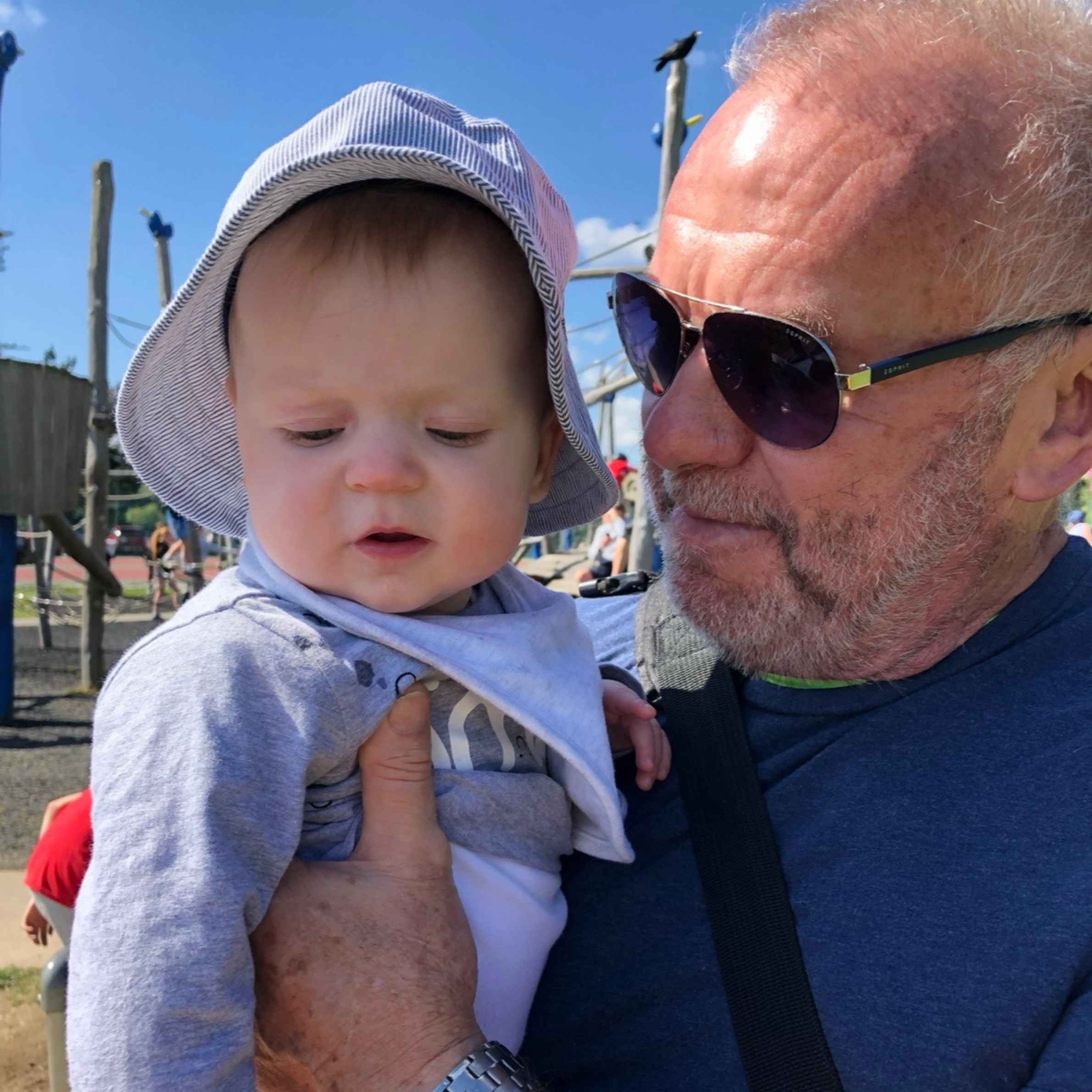Baby wearing grey sunhat and baby grow. Man to the right is holding the baby. He is wearing a navy t-shirt and sunglasses, he is bald. 