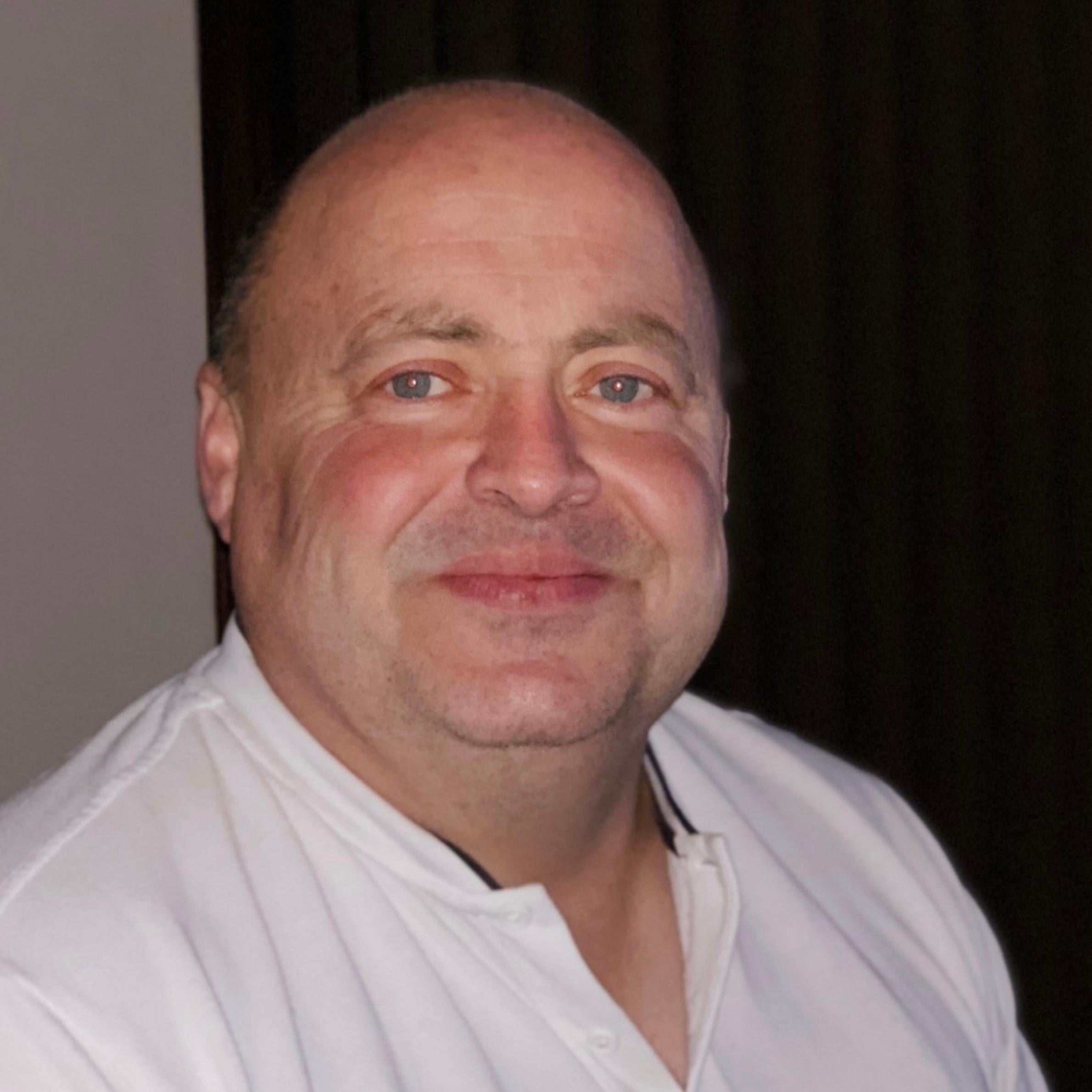 Bald man smiling. He is wearing a white button up top. He has blue eyes.