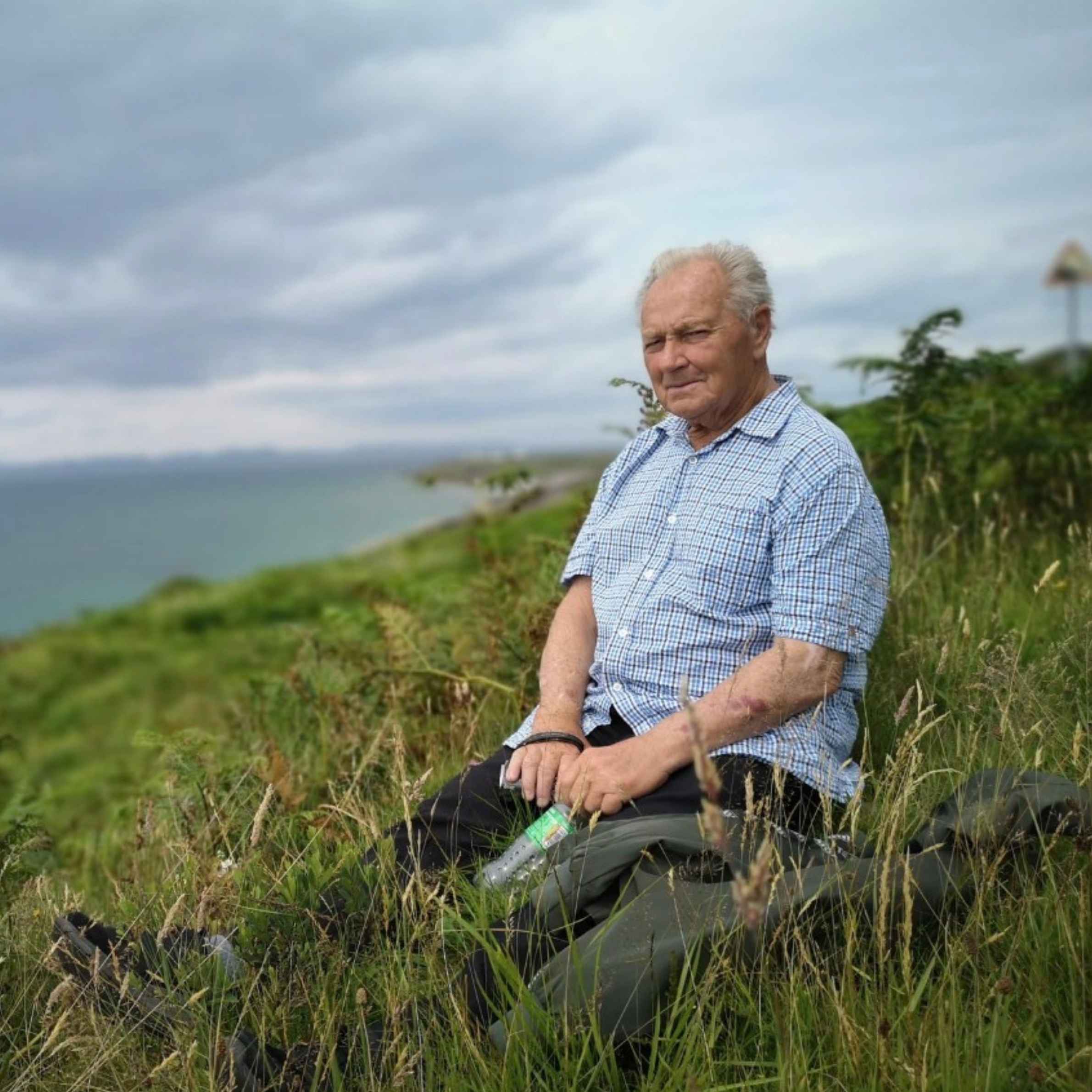 Older man in a blue checkered shirt sits in tall coastal grass beside a walking stick, overlooking the sea beneath a cloudy sky, calm.