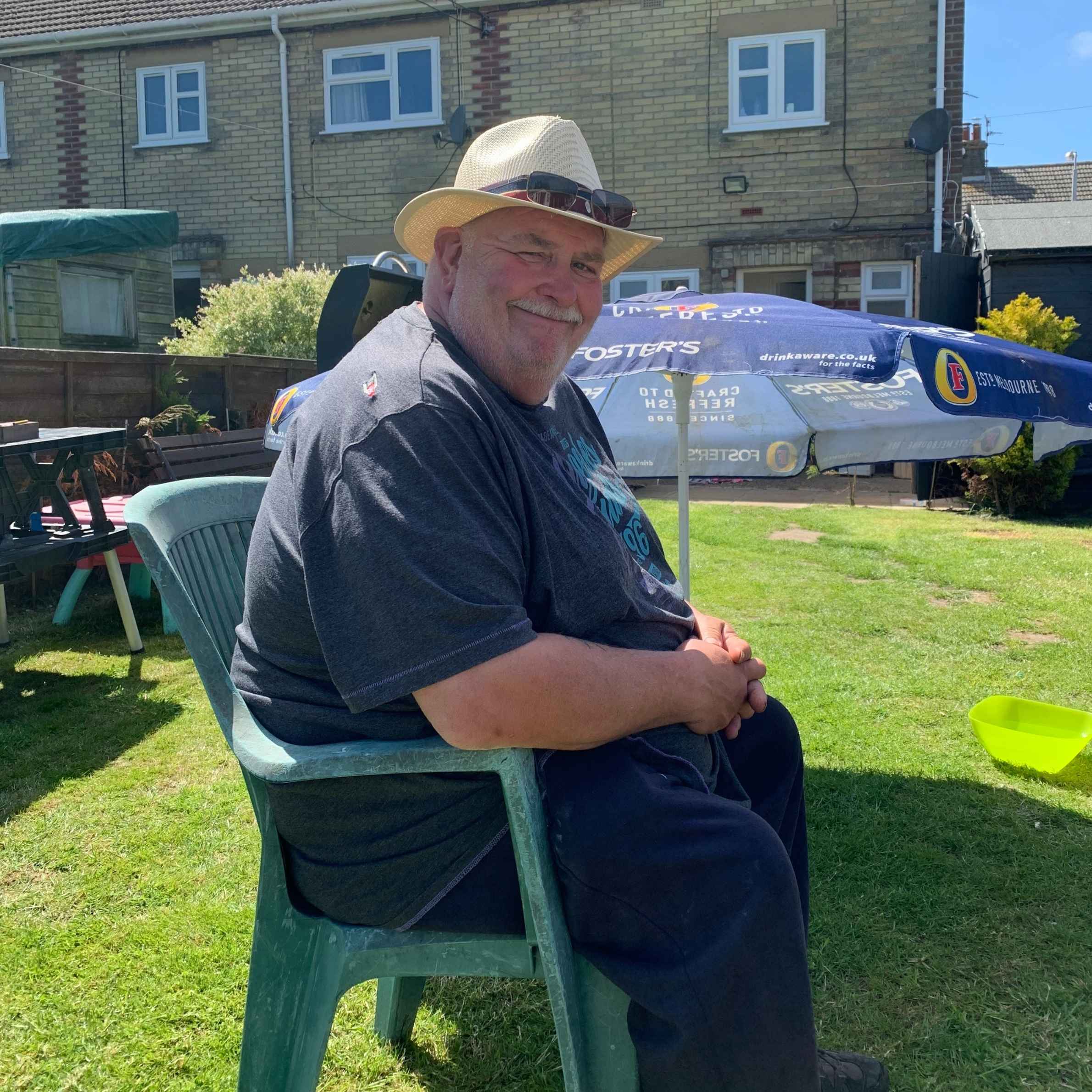 Man sat on green deck chair, he is wearing a short sleeved navy shirt and a cream fedora sunhat. In the background is a navy sun umbrella and in the background is a house. 