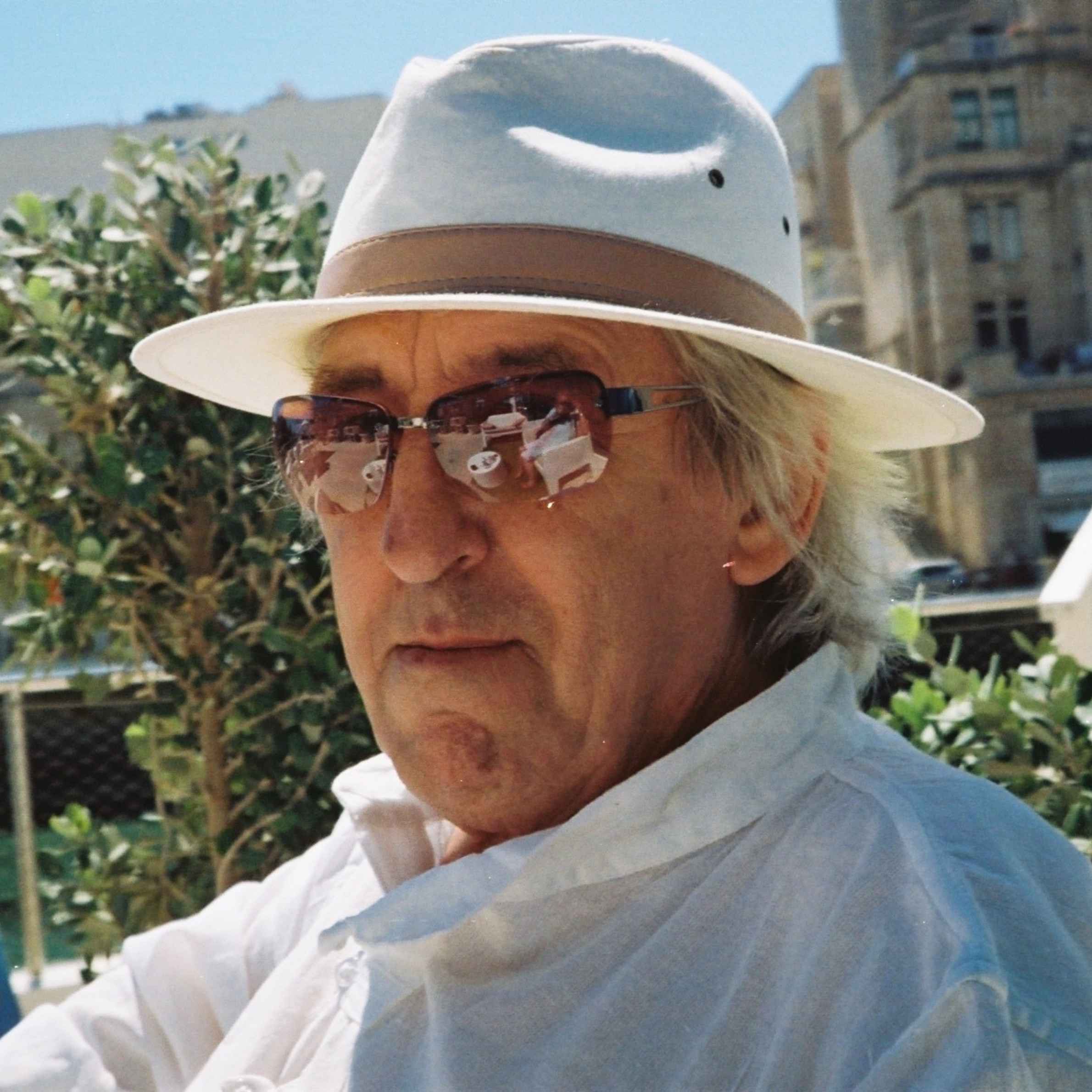 Man wearing white shirt, sunglasses and white fedora hat. He is looking at the camera. In the background there is a green tree and building. 