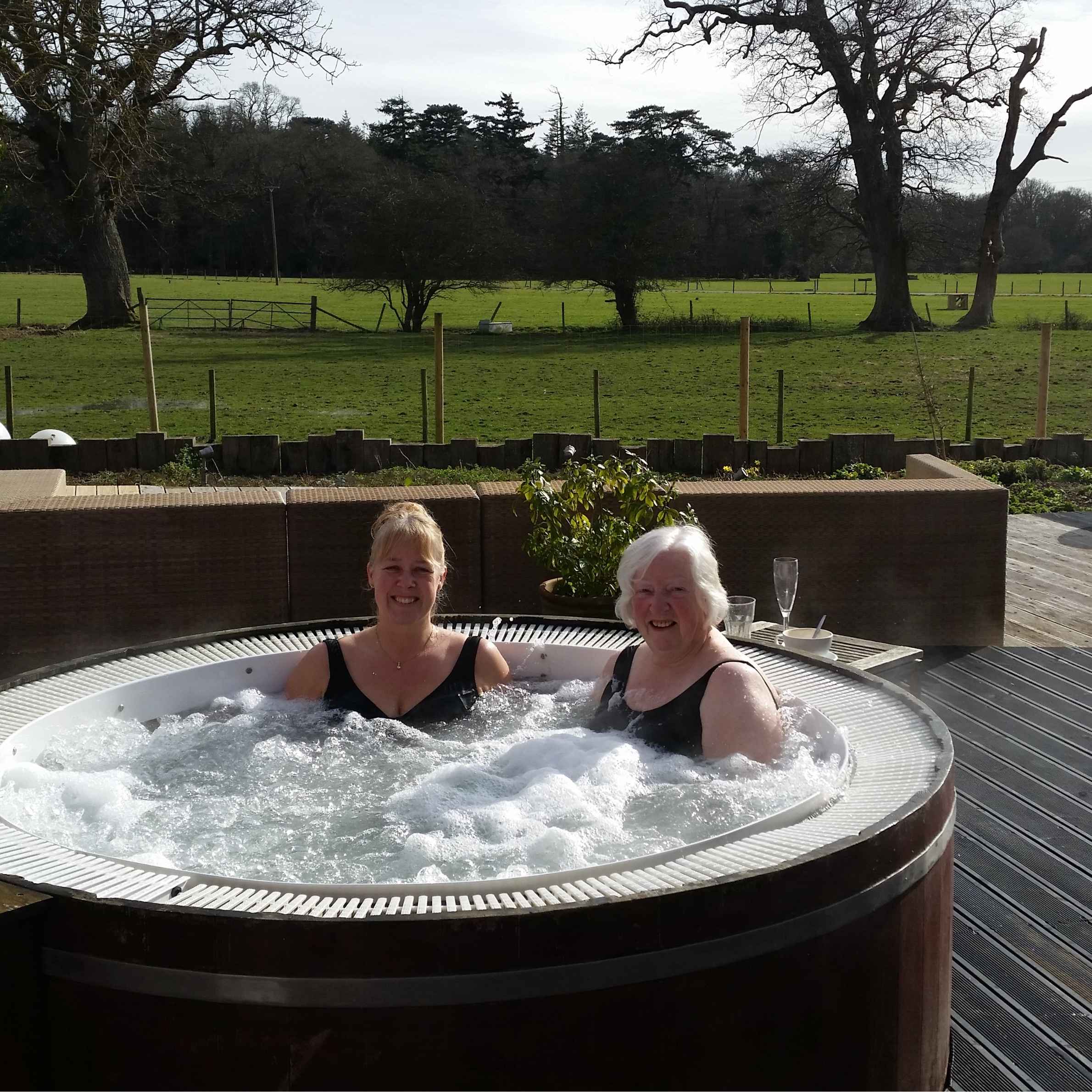 Two ladies sat in a hot tub, looking and smiling at the camera. They are both wearing black swimsuits. IN the background there is a field and trees with wooden decking. 