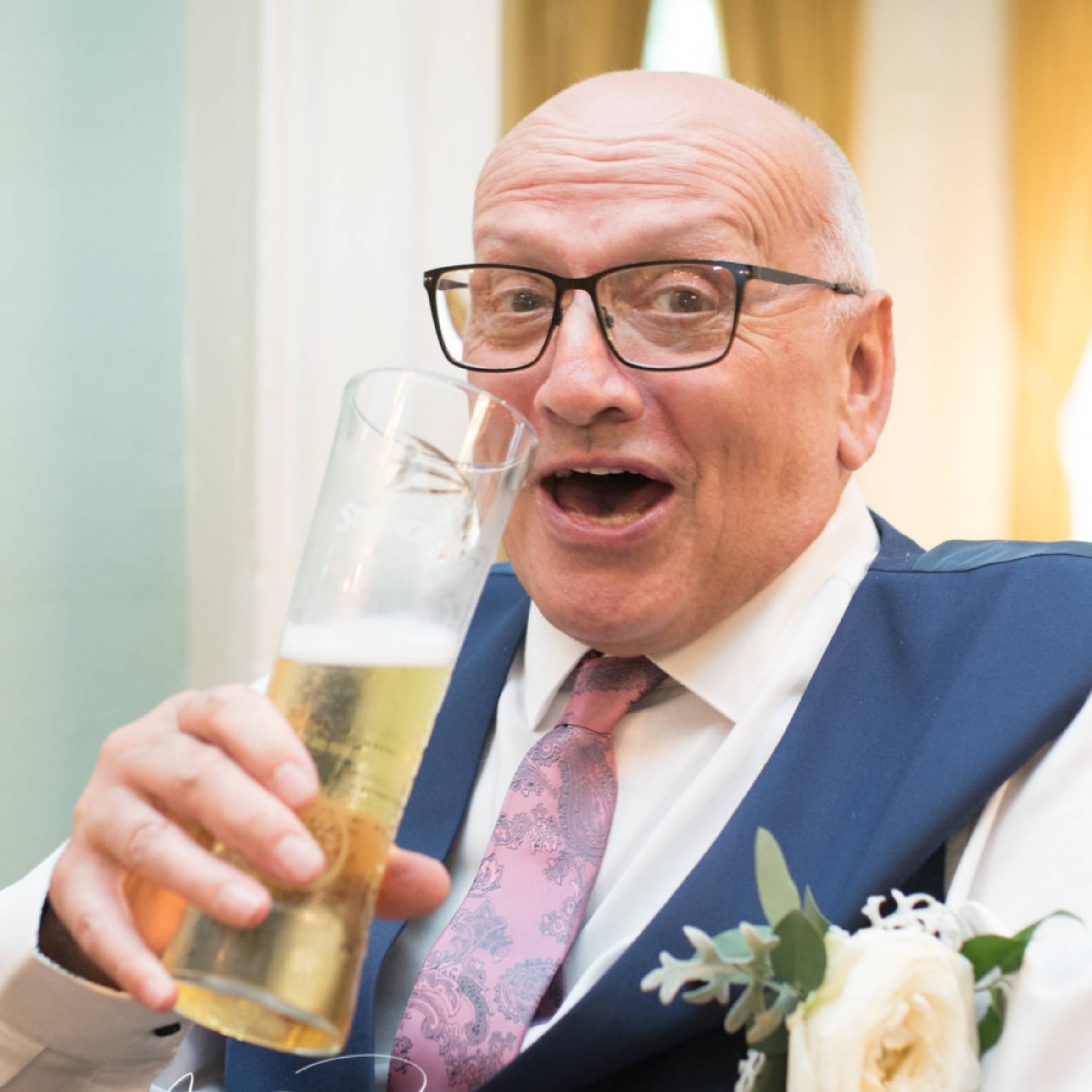 Man wearing white shirt, navy waistcoat and pink tie. He is wearing glasses and holding a pint of beer. 