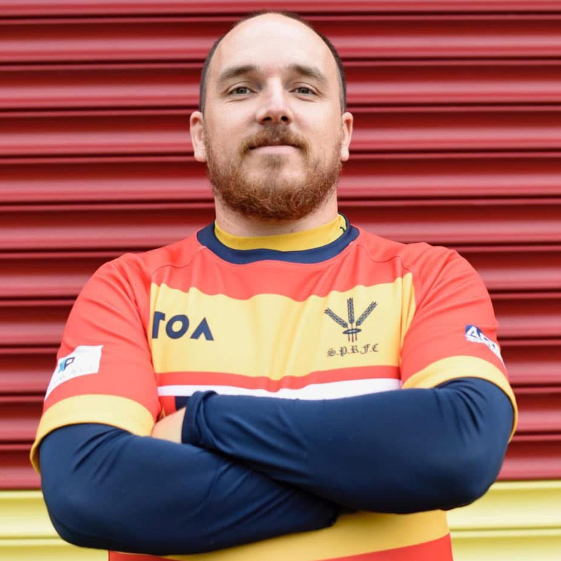 A man is standing with his arms crossed and is wearing a red, yellow, white and blue rugby top. The man has very short hair and has a beard. He is stood in front of red shutters. 