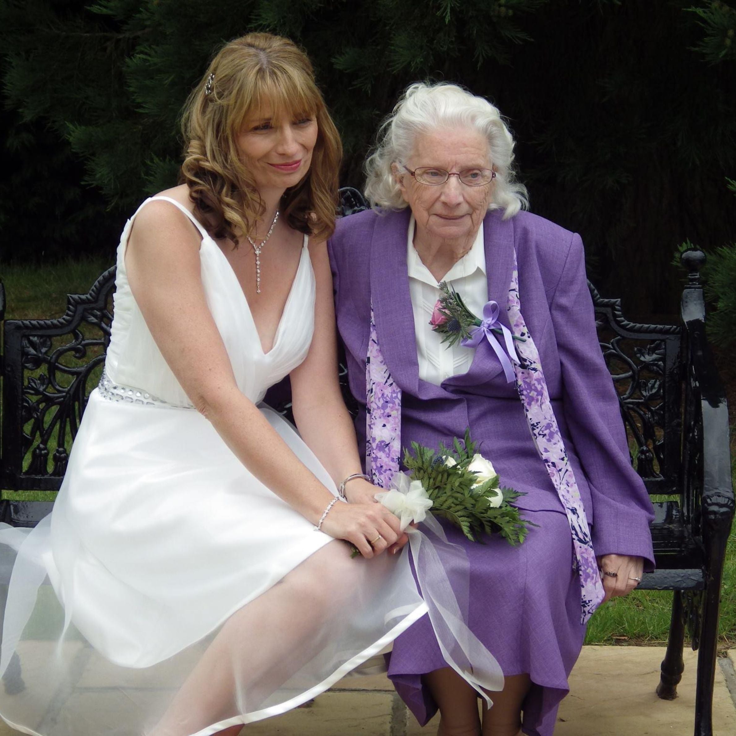 a woman with mid length light brown hair in a white wedding dress holding flowers sat next to an older woman with white hair wearing a purple dress. there is greenary behind them