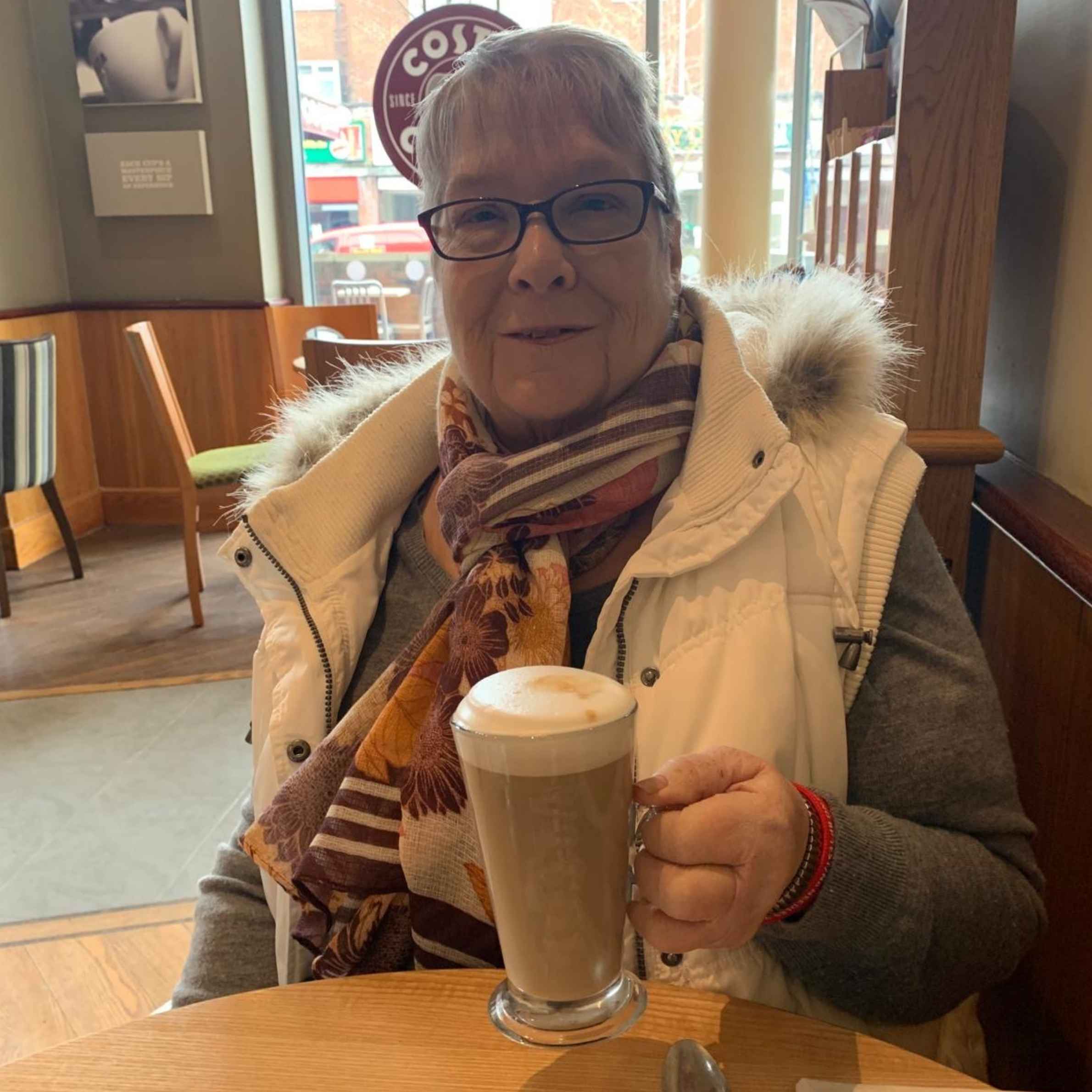 A woman sat in Costa Coffee holding a latte in her right hand. She is wearing a white puffy gilet with a faux fur trim on the hood with a grey top underneath and a scarf