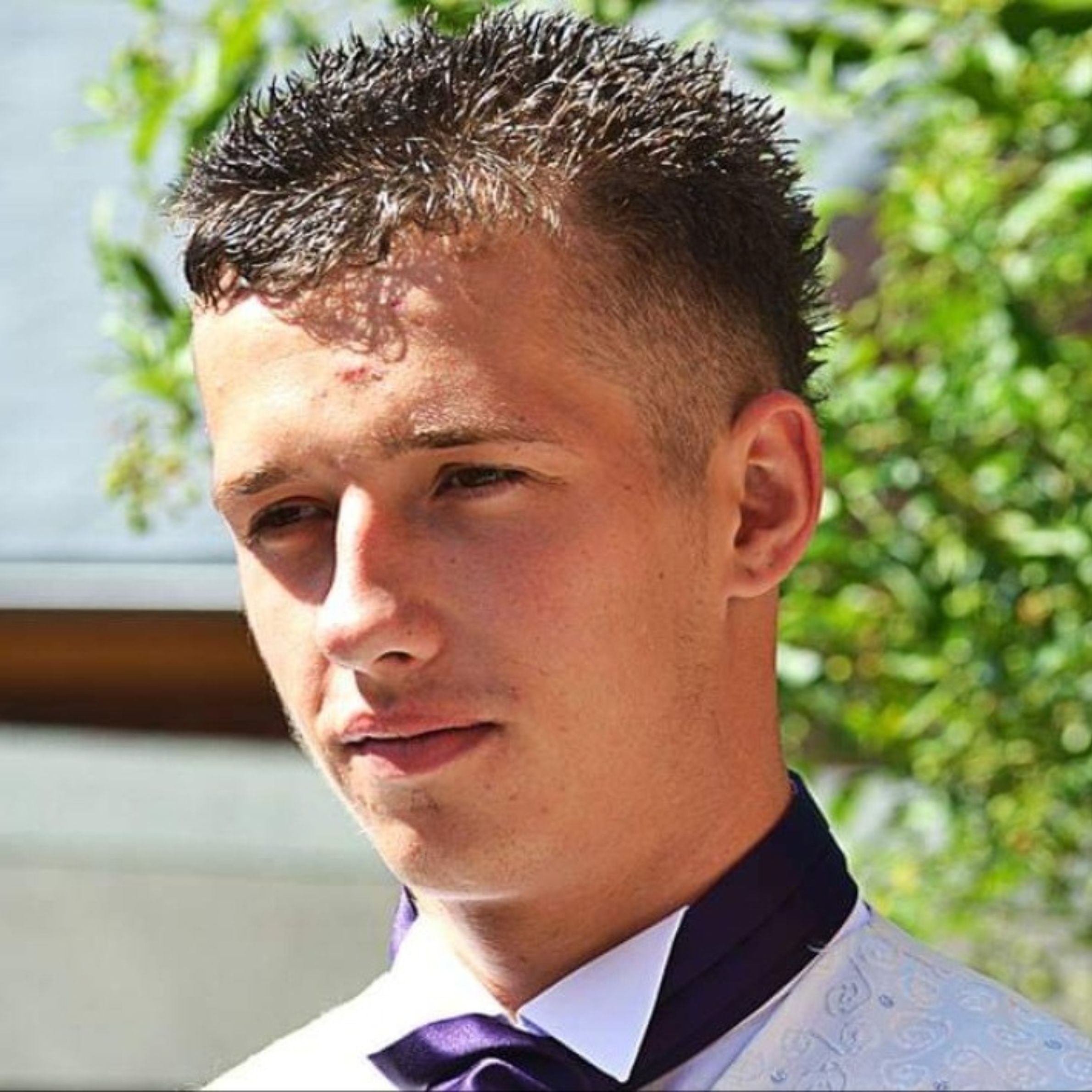 A young man wearing a formal white shirt and a purple tie. It is a sunny day and there are trees in the background.