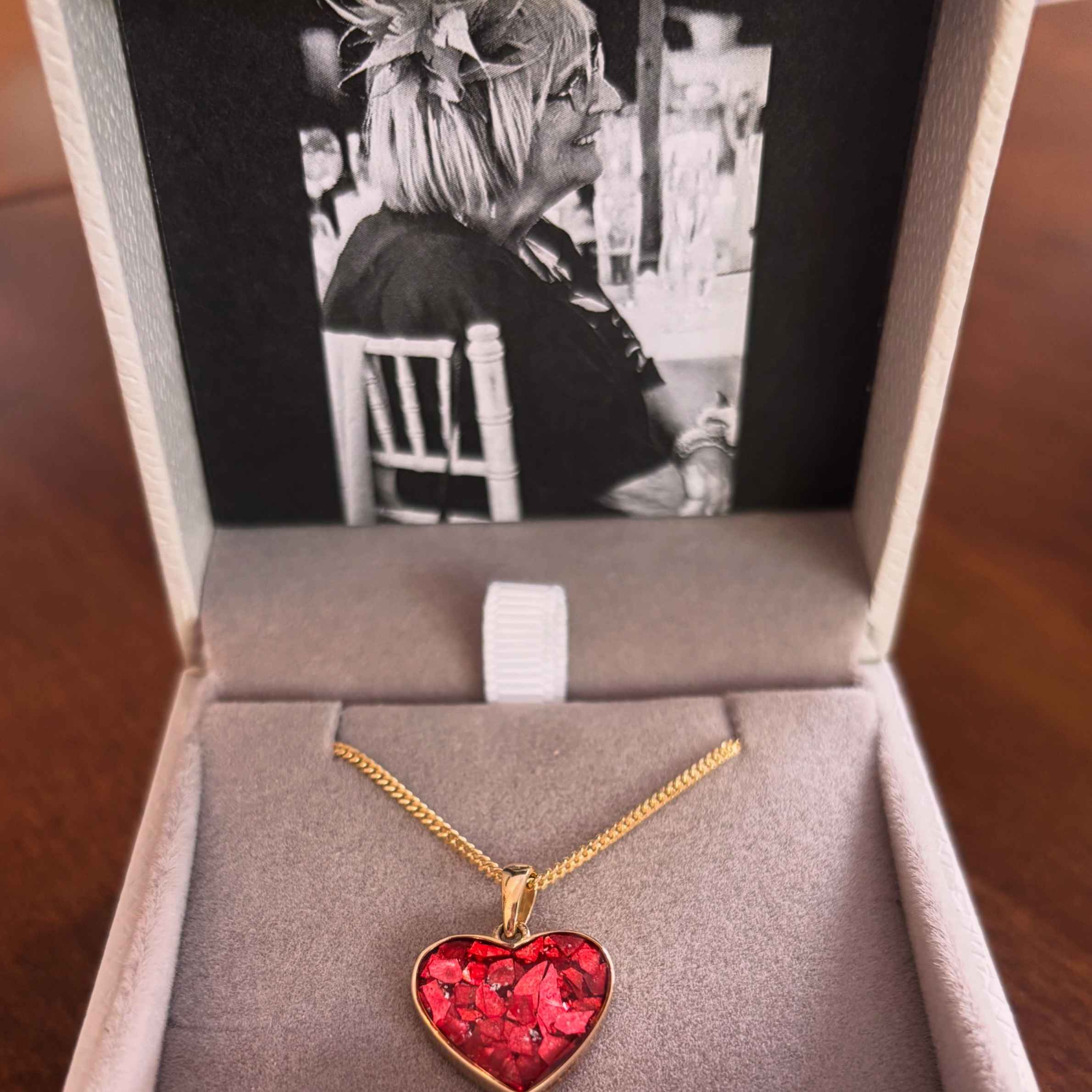 A gold necklace with a red heart charm. The necklace is in a grey jewellery box with a black and white image inside of a woman sat on a chair smiling.