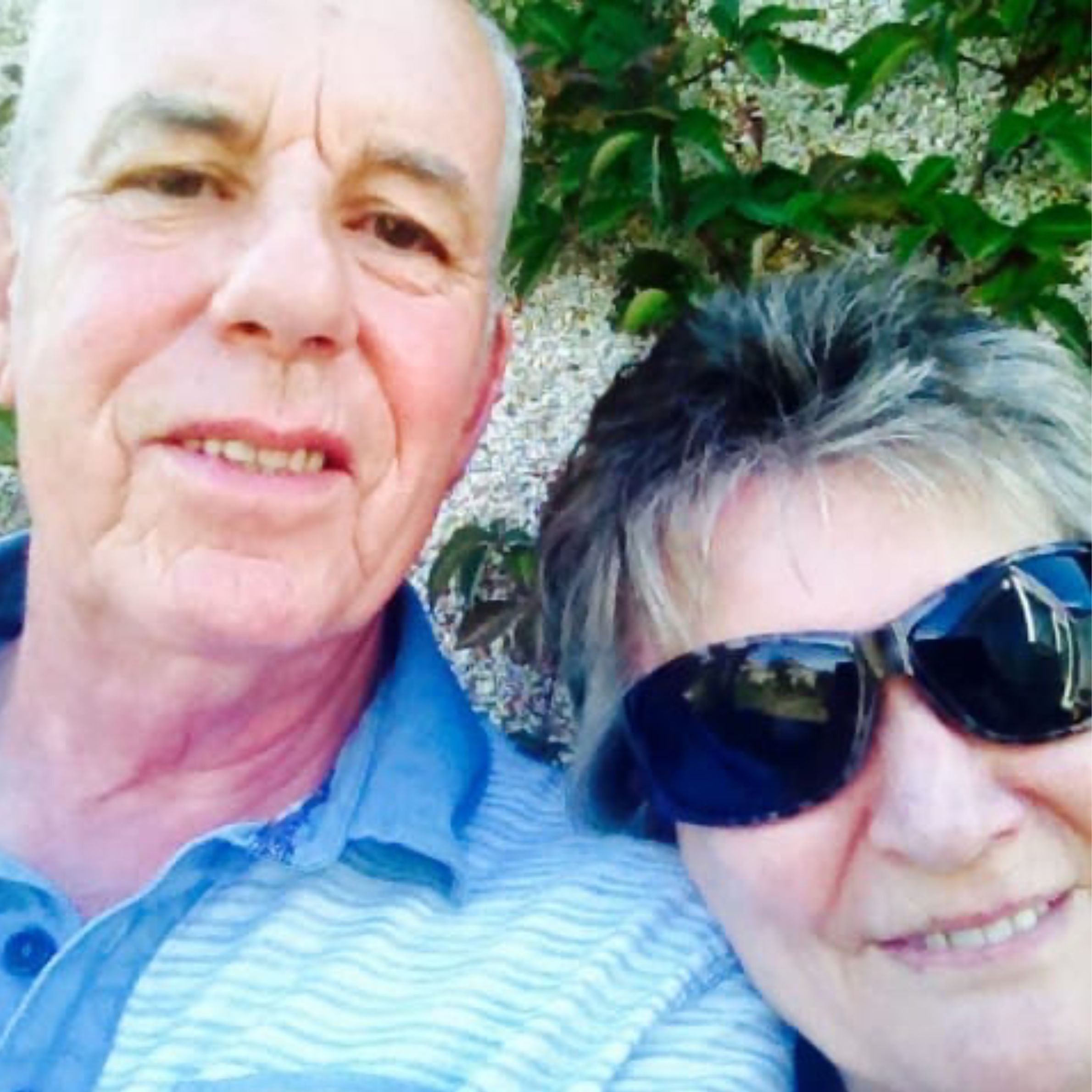An older couple taking a selfie. The gentleman is wearing a striped polo shirt in light and dark blue and the woman has dark sunglasses on.