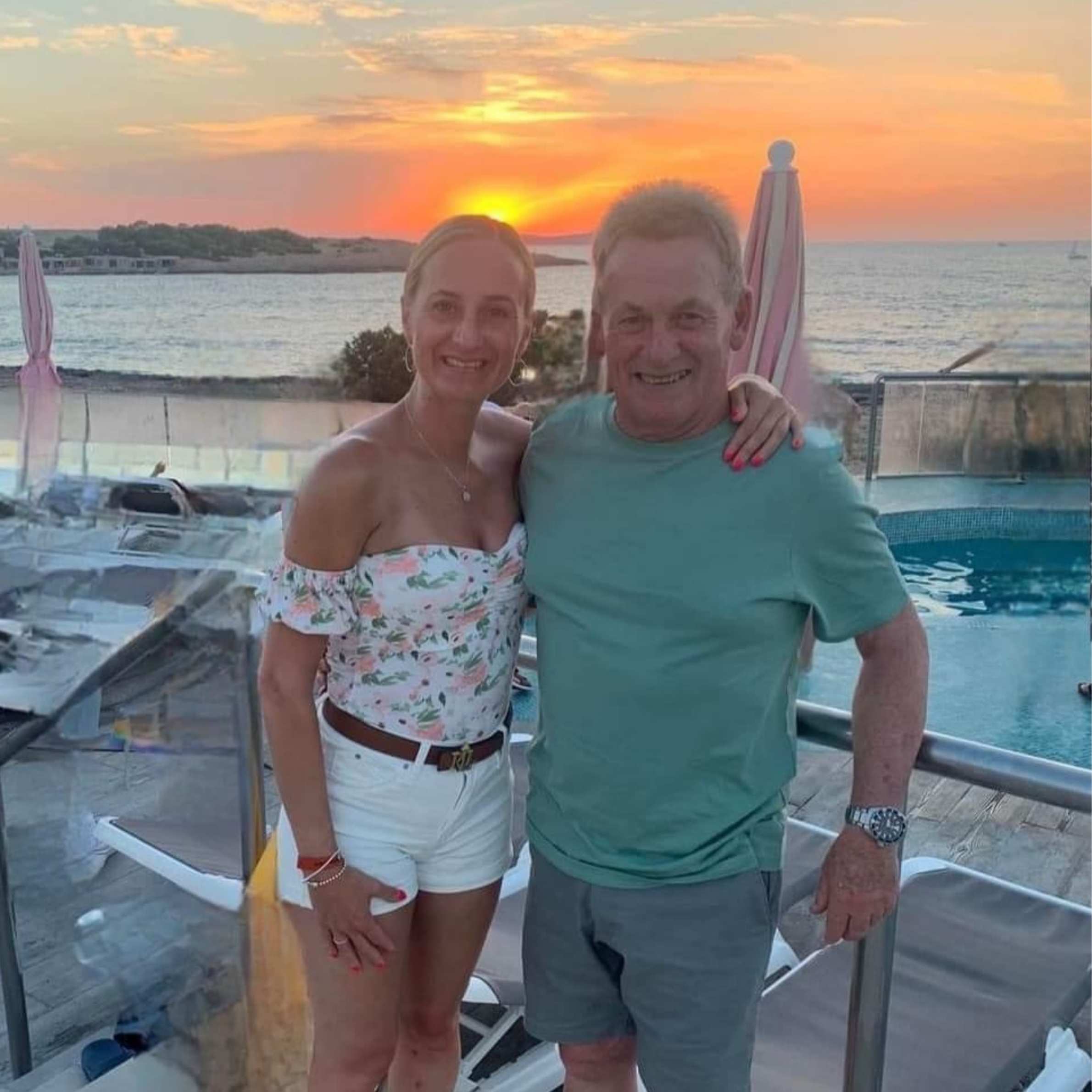 lady stood to the left wearing white shorts and floral white and pink top. to the right is a man wearing a green t-shirt and grey shorts. In the background there is a swimming pool, sunset and sun loungers.