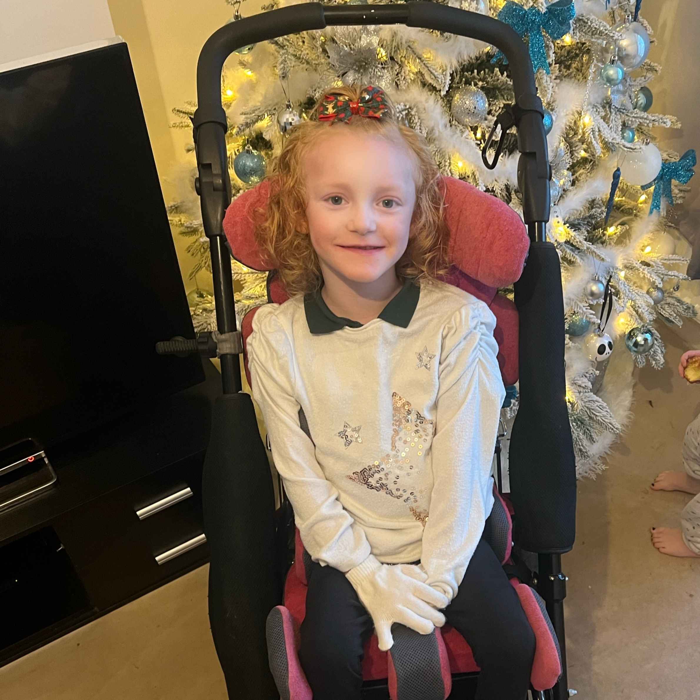 Girl sat in a chair in front of a white Christmas tree. She is wearing a cream jumper with black collared top underneath, and wearing white gloves. She has ginger curly hair. 