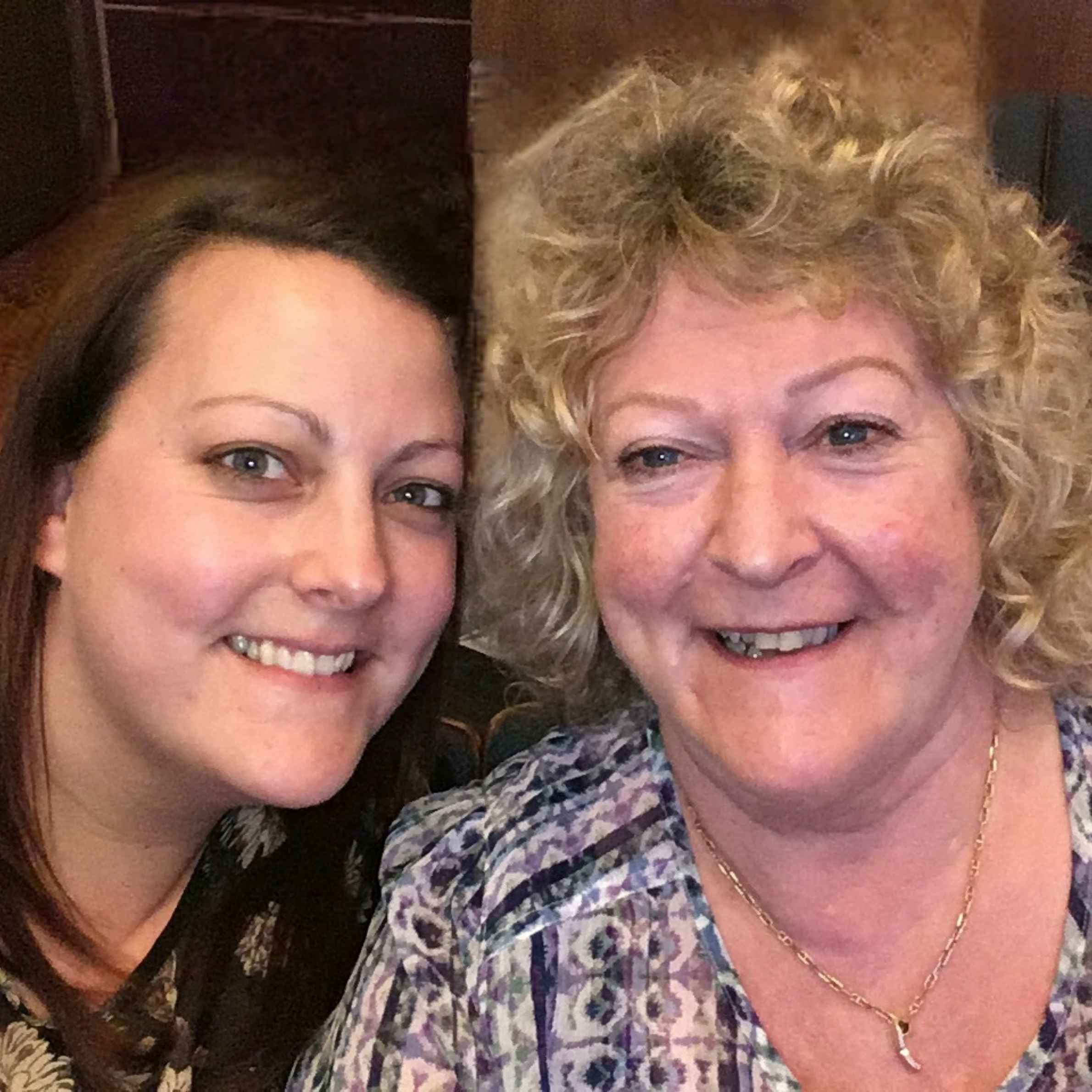 Two ladies smiling. The lady to the left has brown hair and is wearing a dark top, the lady to the right has blonde curly hair, she is wearing a patterned v neck blouse and gold necklace. 