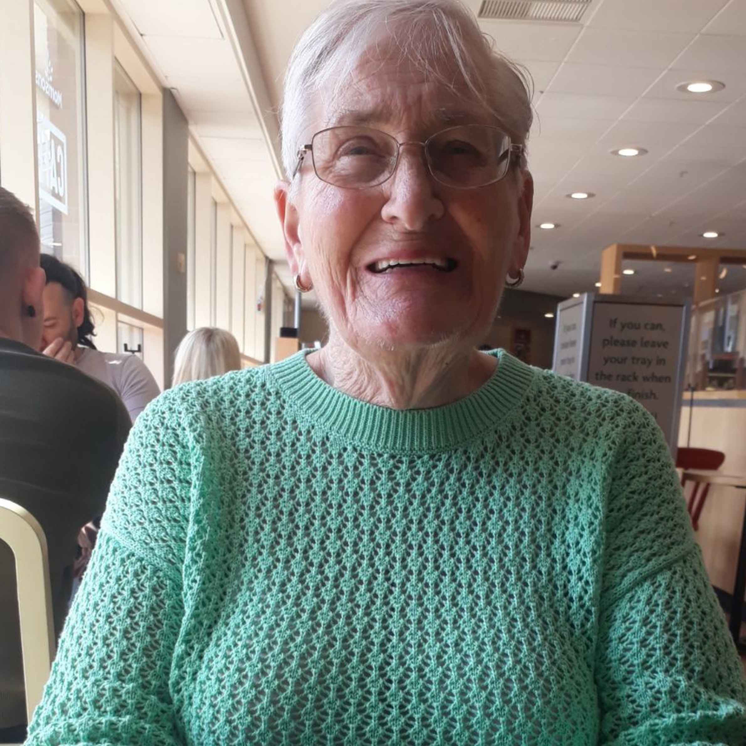Smiling older woman with short gray hair and glasses, wearing a mint-green sweater in a bright, welcoming café or restaurant.