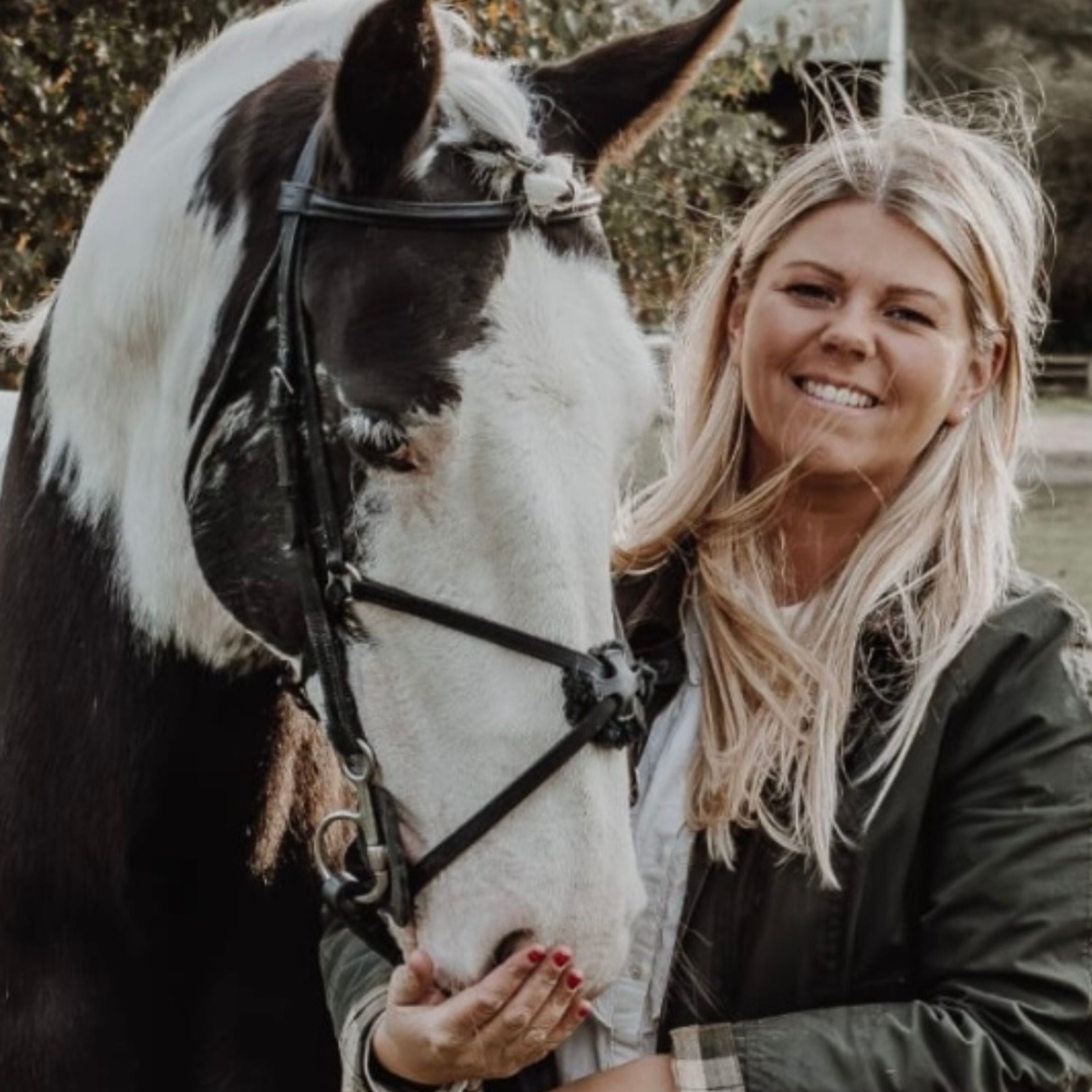 A photo of a woman and a horse. The horse is black and white piebald and is wearing a black bridle. The woman has long blonde hair and is wearing a green jacket. She is smiling and is cupping the horses muzzle with her hands.