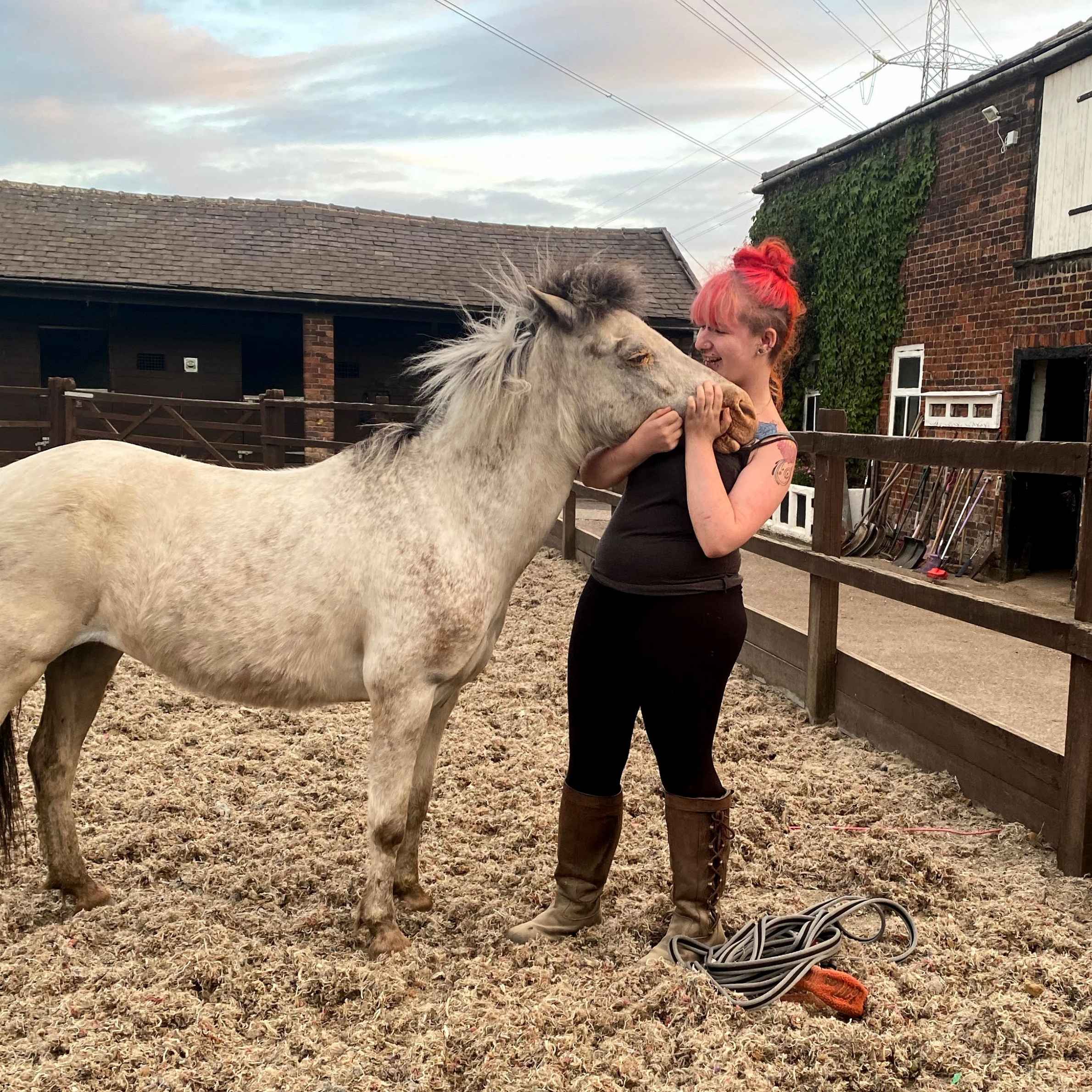A red-haired woman affectionately nuzzles a small gray horse in a rustic stable yard, conveying warmth, trust, and companionship.