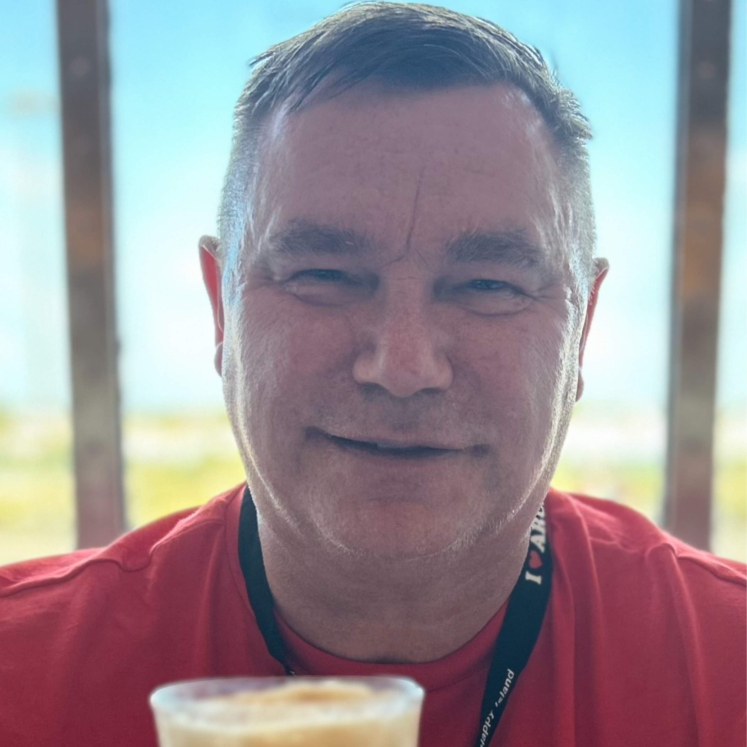 A man wearing a red tshirt is smiling for the camera. He is infront of large windows and it is sunny in the background. He is wearing a black lanyard.
