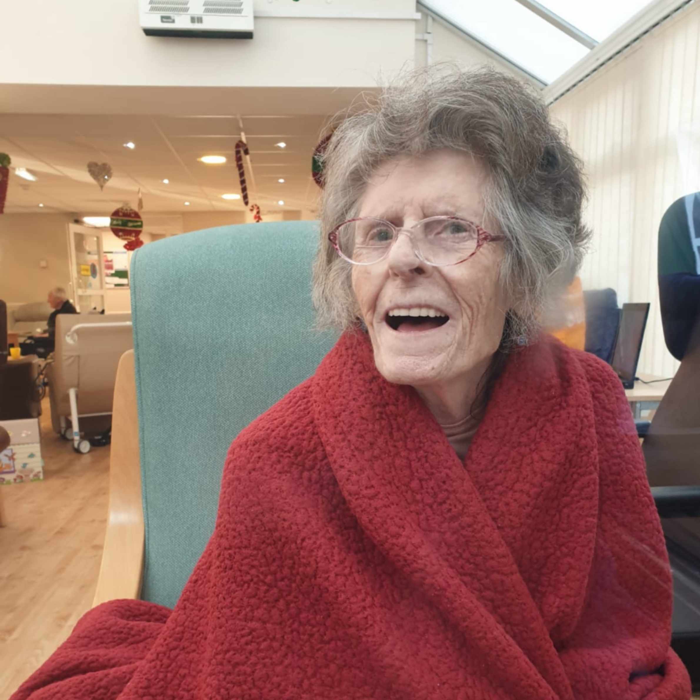 Lady sat on grey backed wooden chair, she is wrapped in a red blanket, smiling and wearing glasses. 