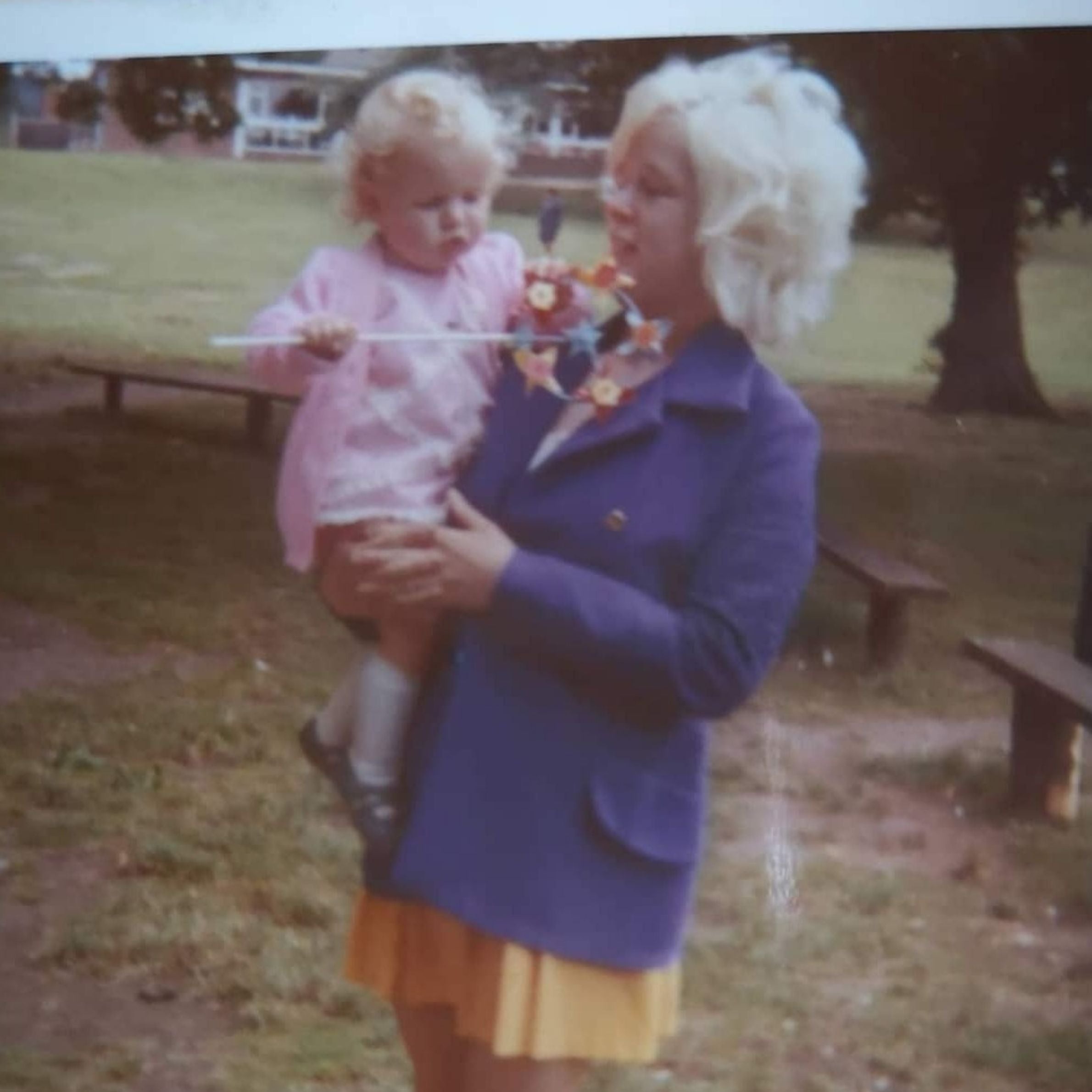 Lady with navy coat on and yellow dress underneath holding little girl. The lady has white short curly hair and the little girl is wearing a pink dress and cardigan and also has white curly hair 