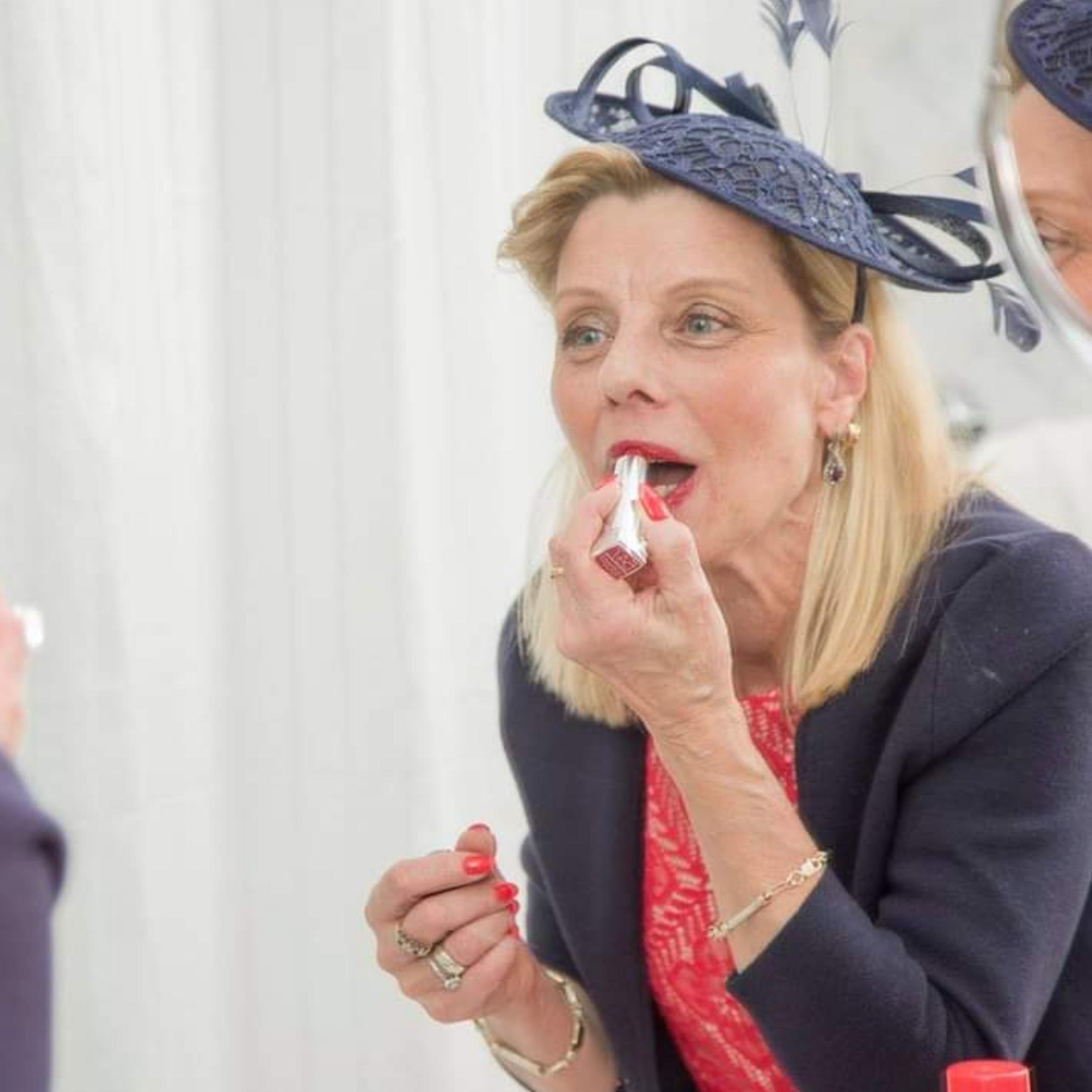 Picture of lady looking into mirror whilst applying red lipstick. She is wearing a navy fascinator with a red dress and navy blazer. She has mid-length blonde hair.