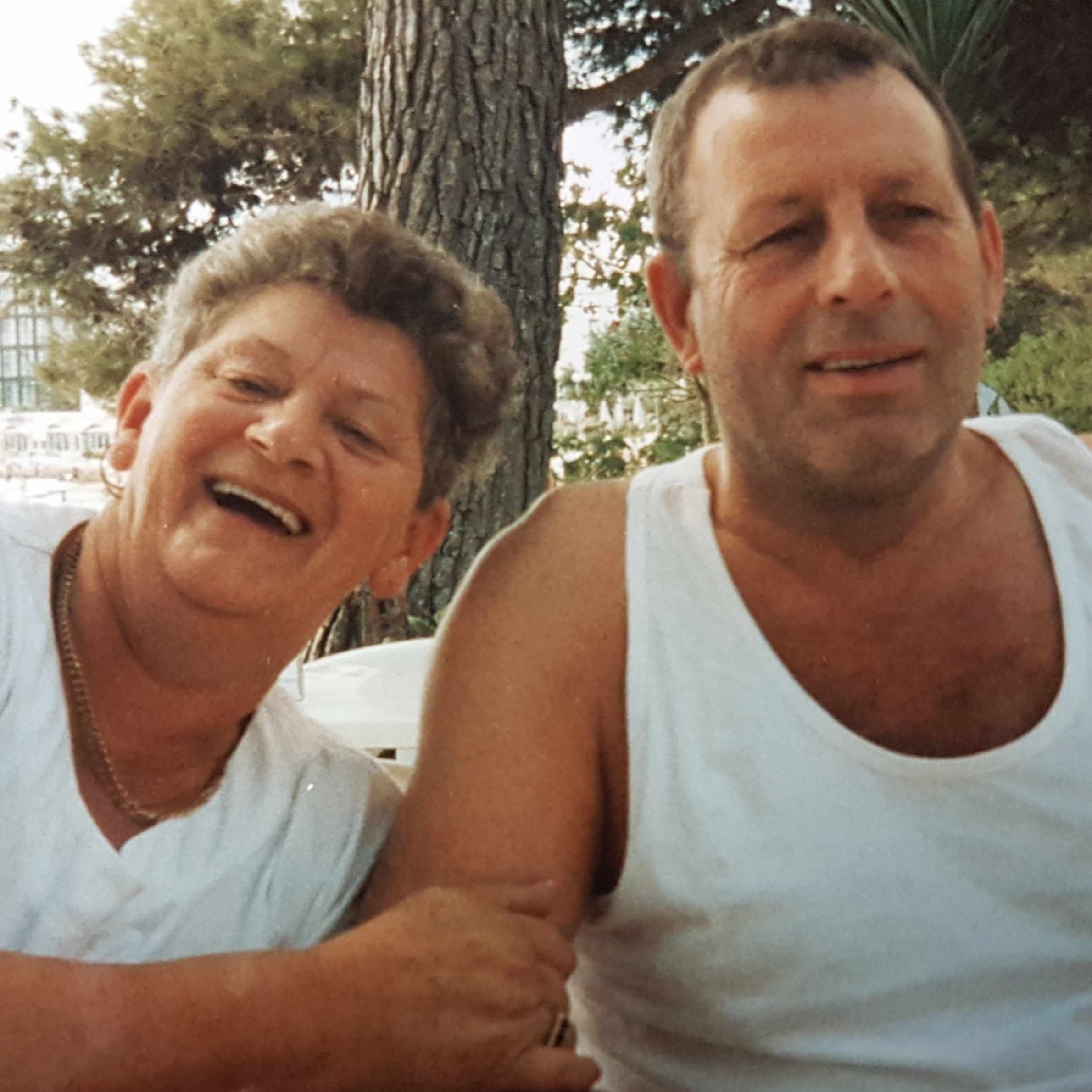 Lady to the left of the photo smiling wearing a white tshirt, she has her arms around the arm of the man to the right of the photo. He is wearing a white vest. There is a tree in the background. They both have brown hair. 