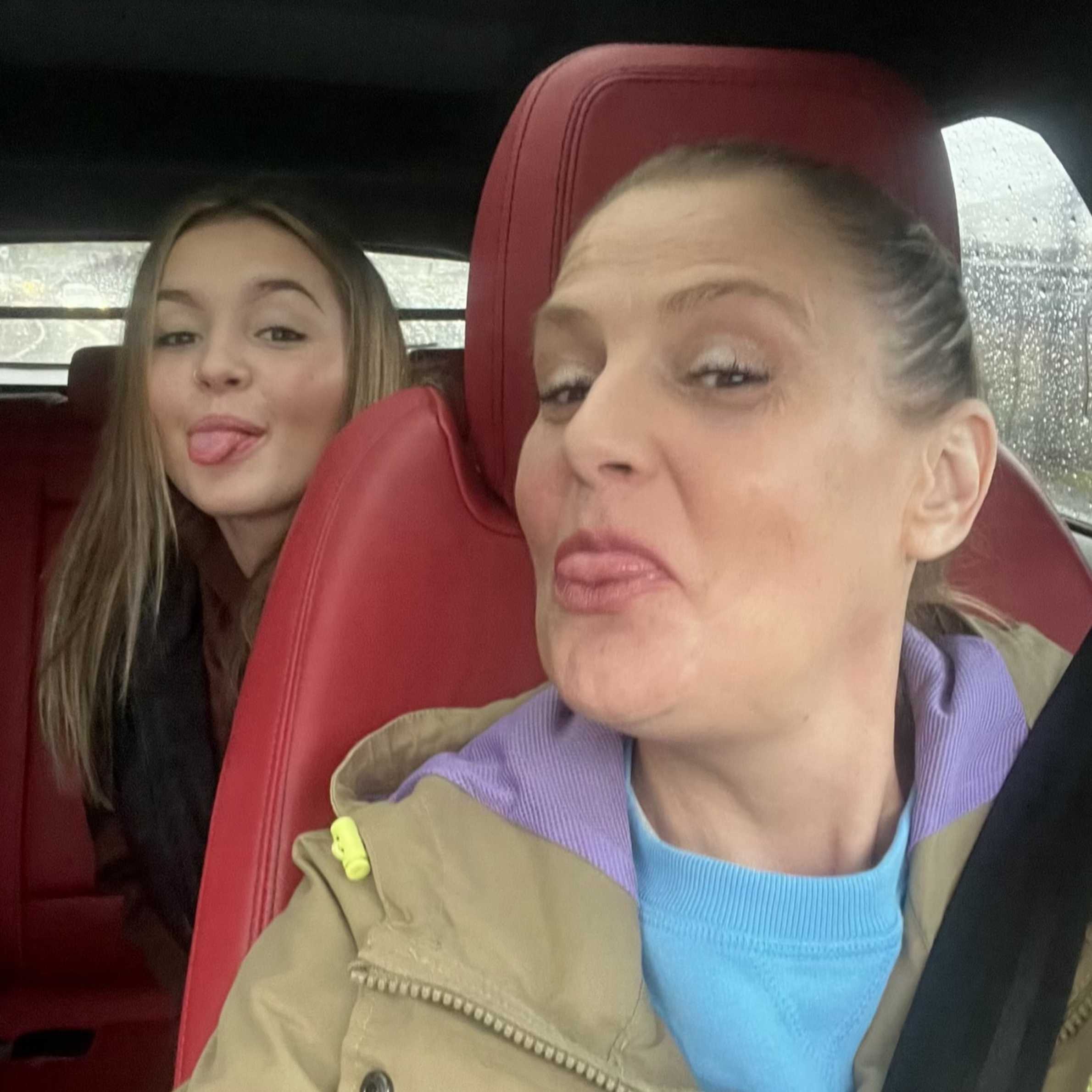 Two women in a car make playful faces for a selfie; the foreground woman wears a tan jacket and blue sweater, while rain dots the window.