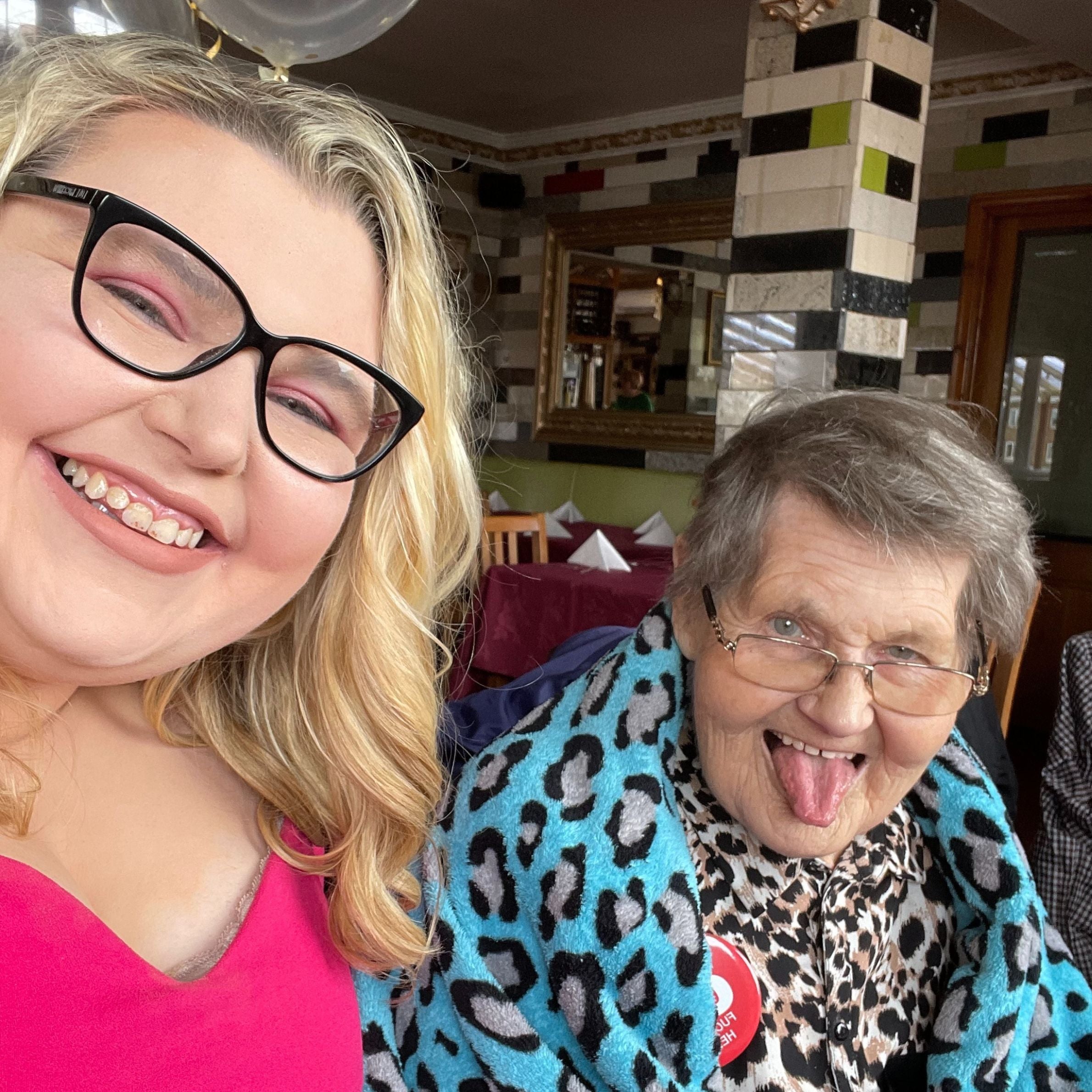 Two women are sat on a chair smiling. One woman has glasses, blonde hair and is wearing a bright pink top. The other woman is sticking her tongue out and is wearing a leopard print top and has a blue leopard print blanket around her shoulders.