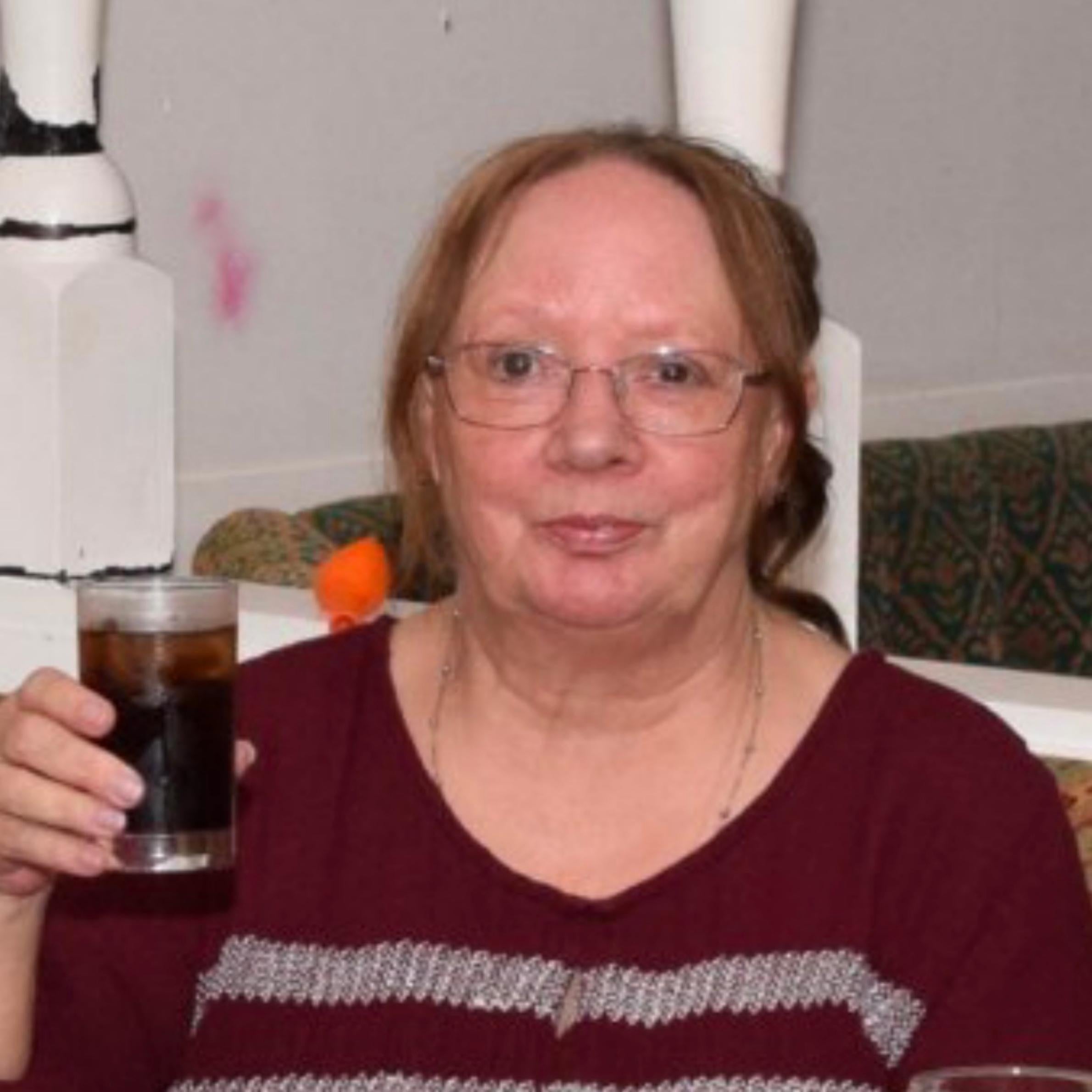  Lady is smiling at the camera, she is holding a drink in her hand. She is wearing a maroon coloured v-neck top with horizontal grey stripes across the front. She is wearing glasses and has auburn coloured hair, tied back.