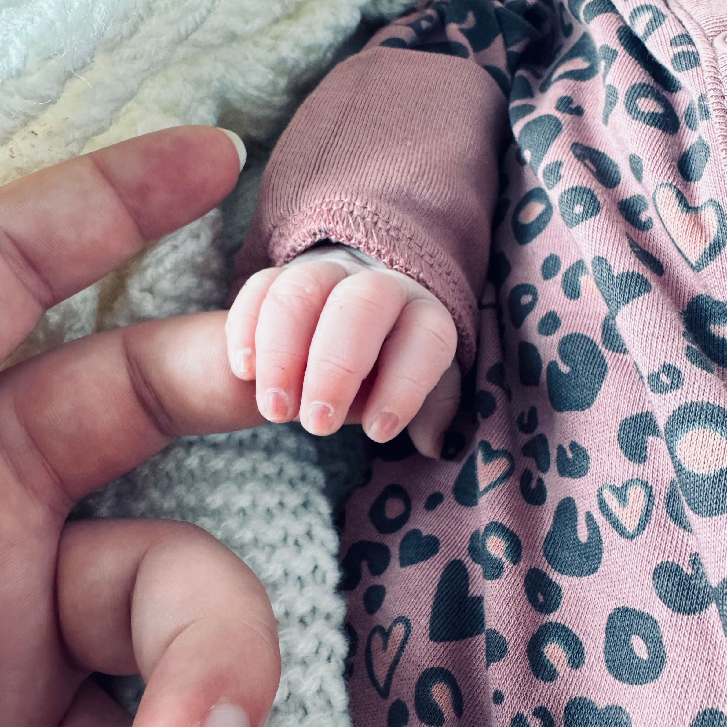 a baby's hand holding an adult finger. the baby is wearing a pink top with black hearts pattered on it.