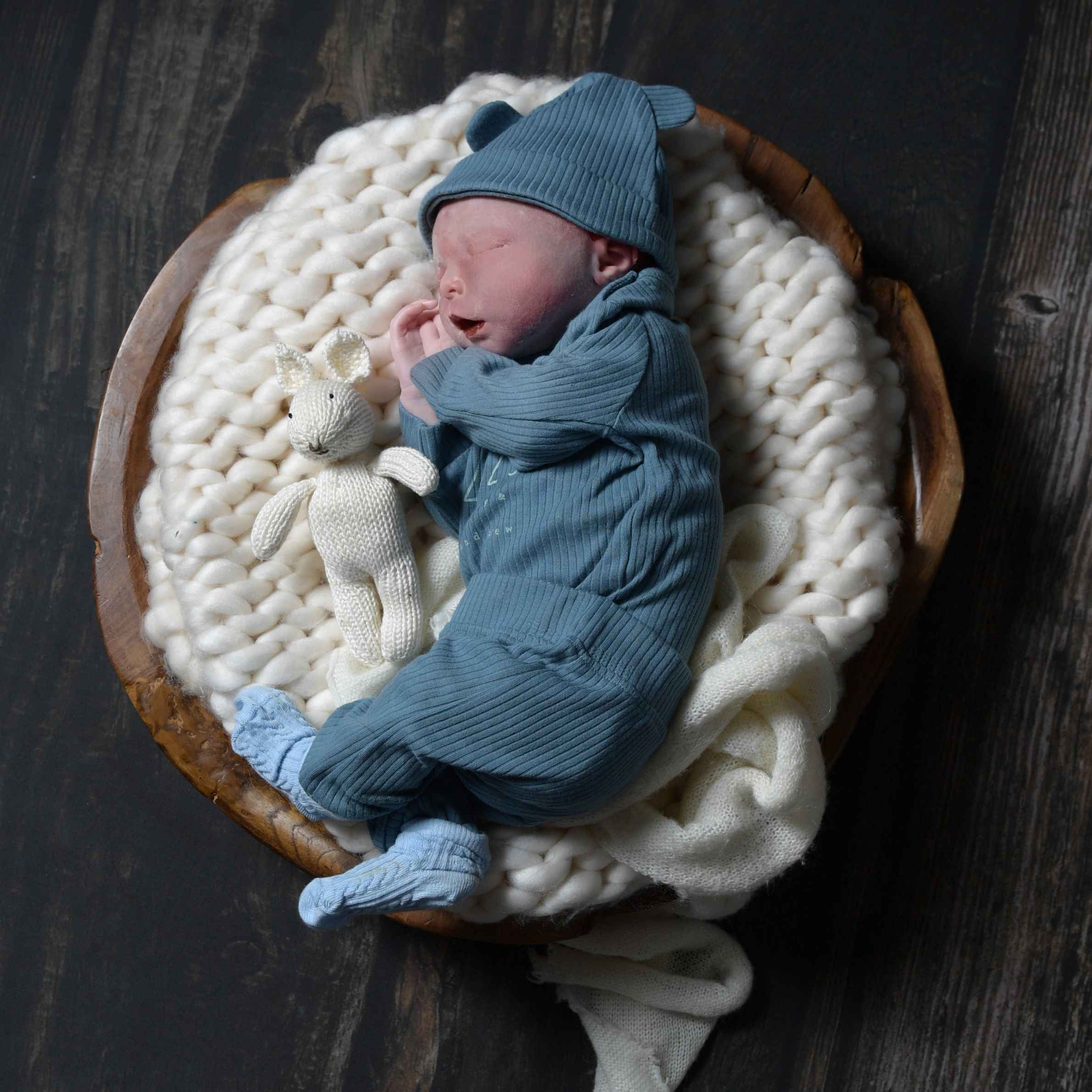 Baby wearing blue sleepsuit and hat, laid in a brown basket on a white knitted blanket. There is a small white bear next to him. 