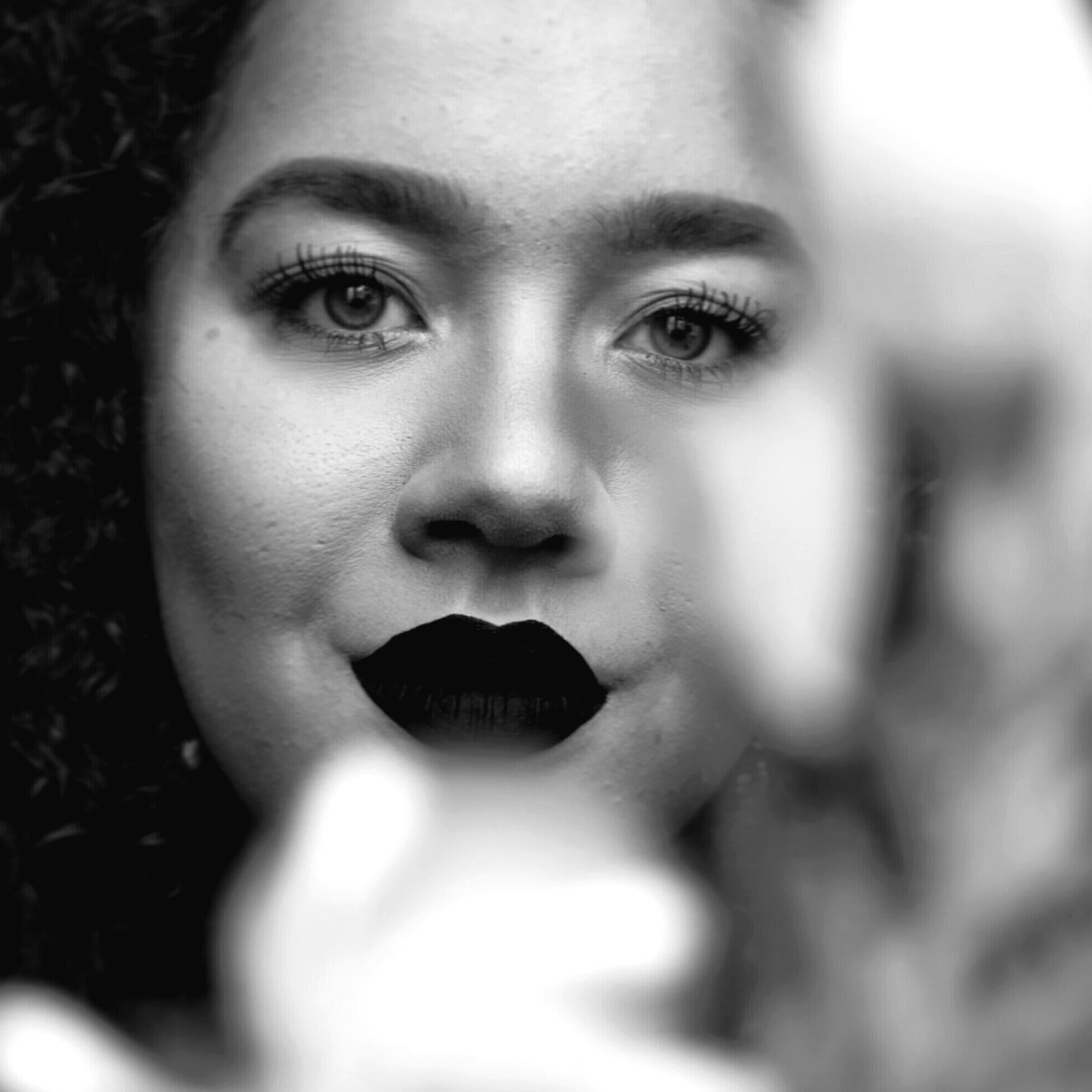 A black and white image of a young lady facing the camera. She is wearing dark lipstick and has large full eyelashes. She is facing towards the camer while an object is out of focus in the foreground
