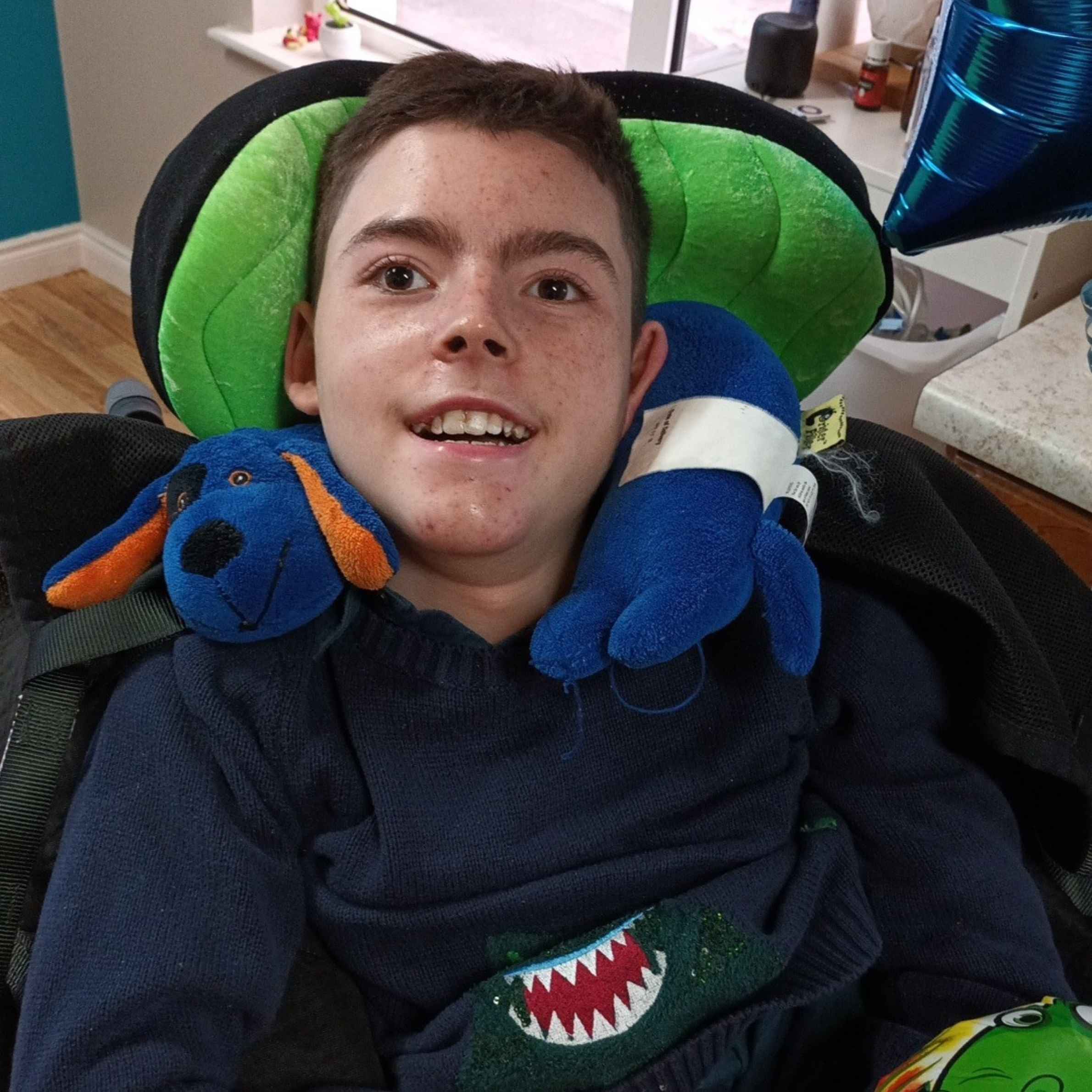 Man sat in green and black chair, he is smiling and looking at the camera. He has brown hair, he has a blue dog teddy rapped around his neck. 