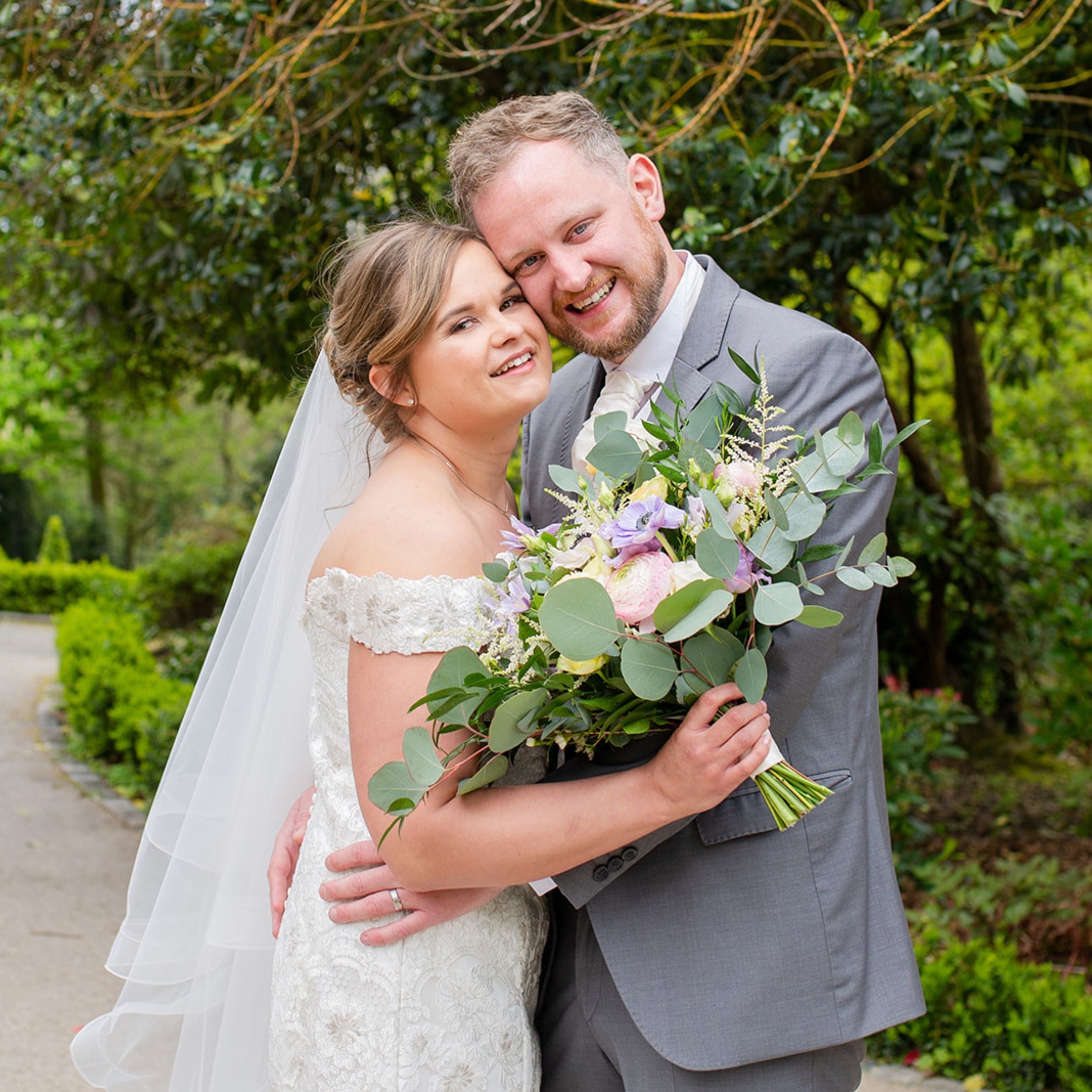 woman in a white wedding dress hugging her new husband next to her who is wearing a grey suit. she is holding some green and purple flowers and they are both smiling. the background is a green garden with trees and a concrete path