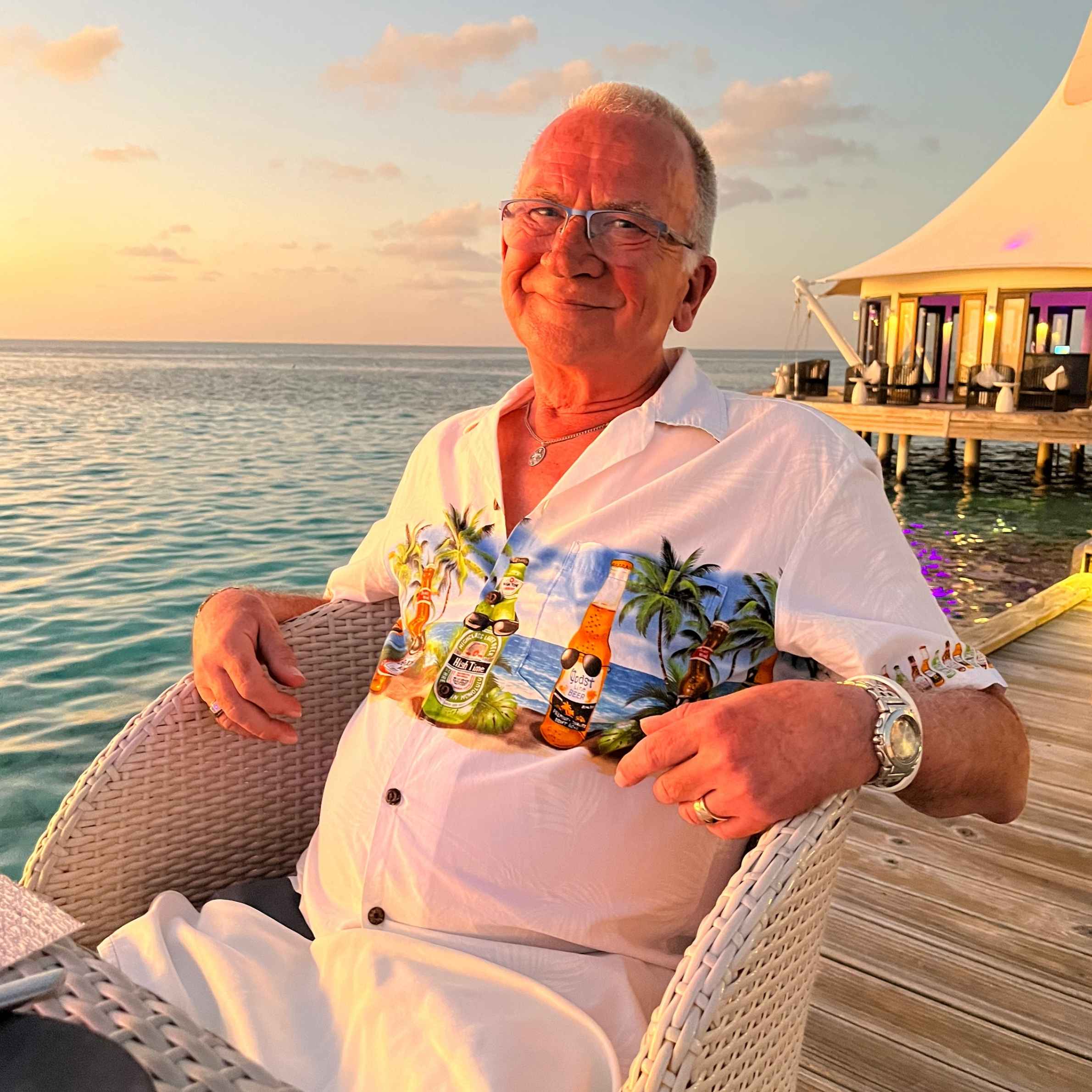 Smiling older man in a colorful floral shirt relaxes in a wicker chair with drinks at a tropical seaside resort during sunset.