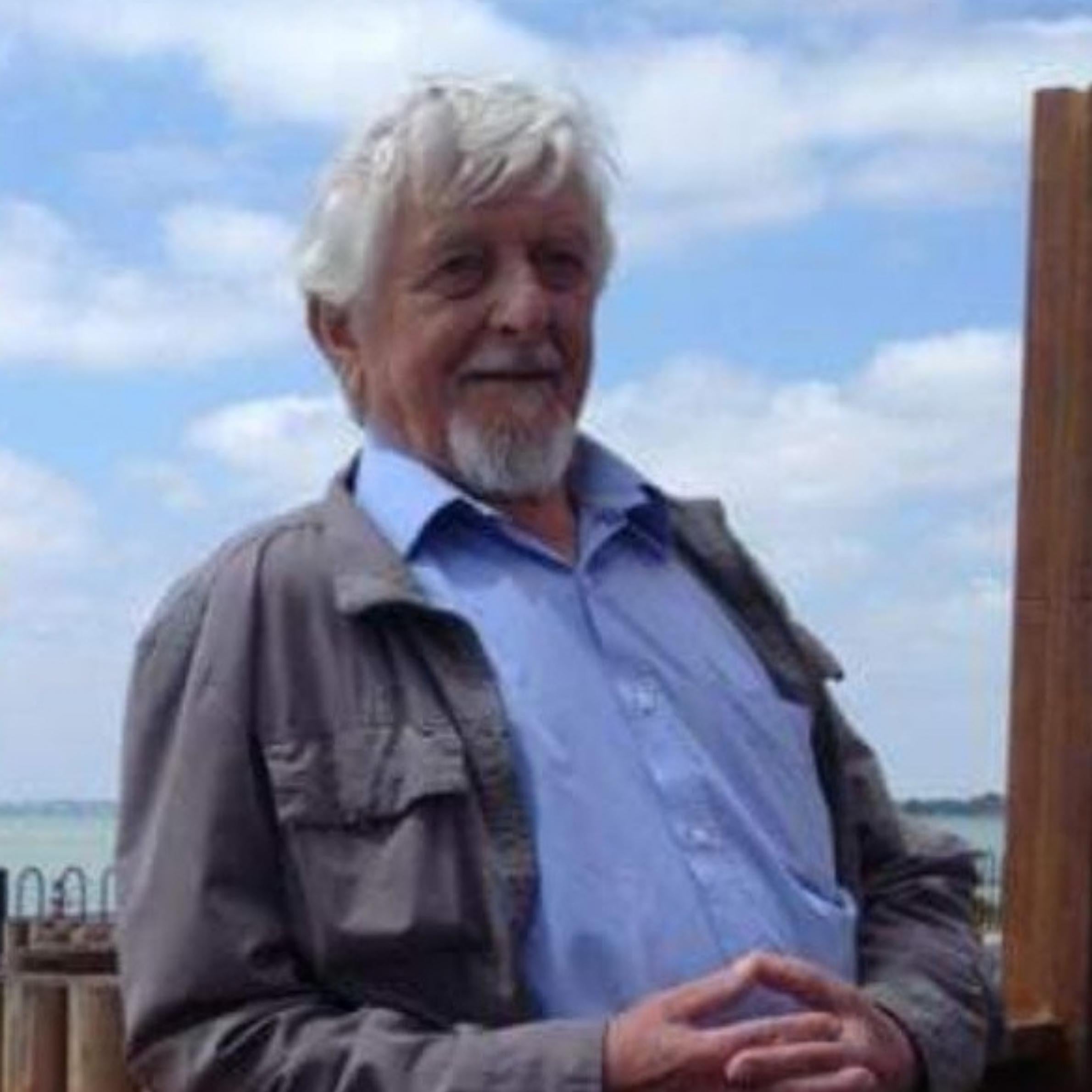 Man waring grey jacket with blue shirt underneath. He has grey hair and a grey beard. Behind him is the blue sky. 
