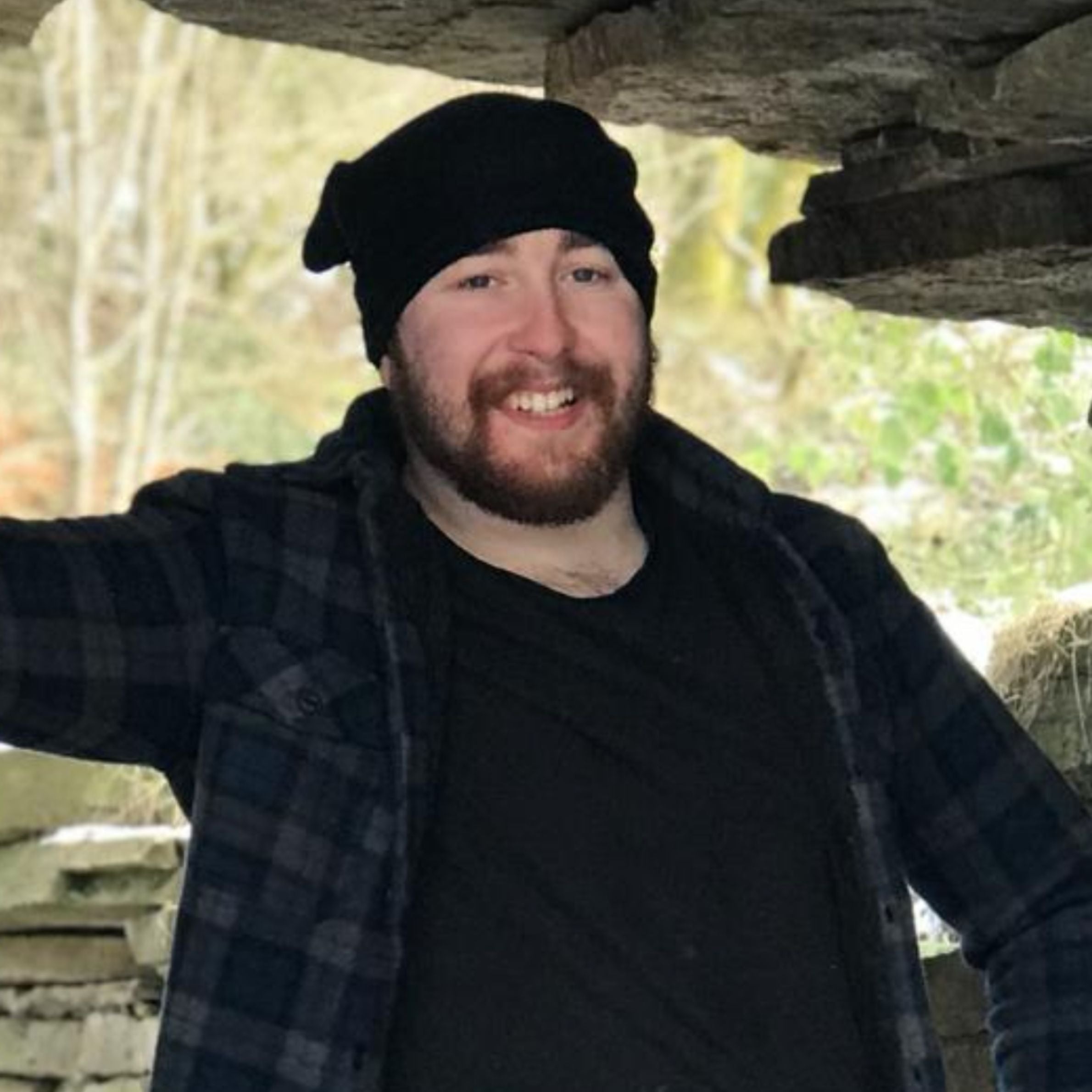 man wearing a dark blue checkered shirt with a black t-shirt underneath. he is also wearing a black hat and is smiling and has a dark hair beard. there is rocks and trees in the background 