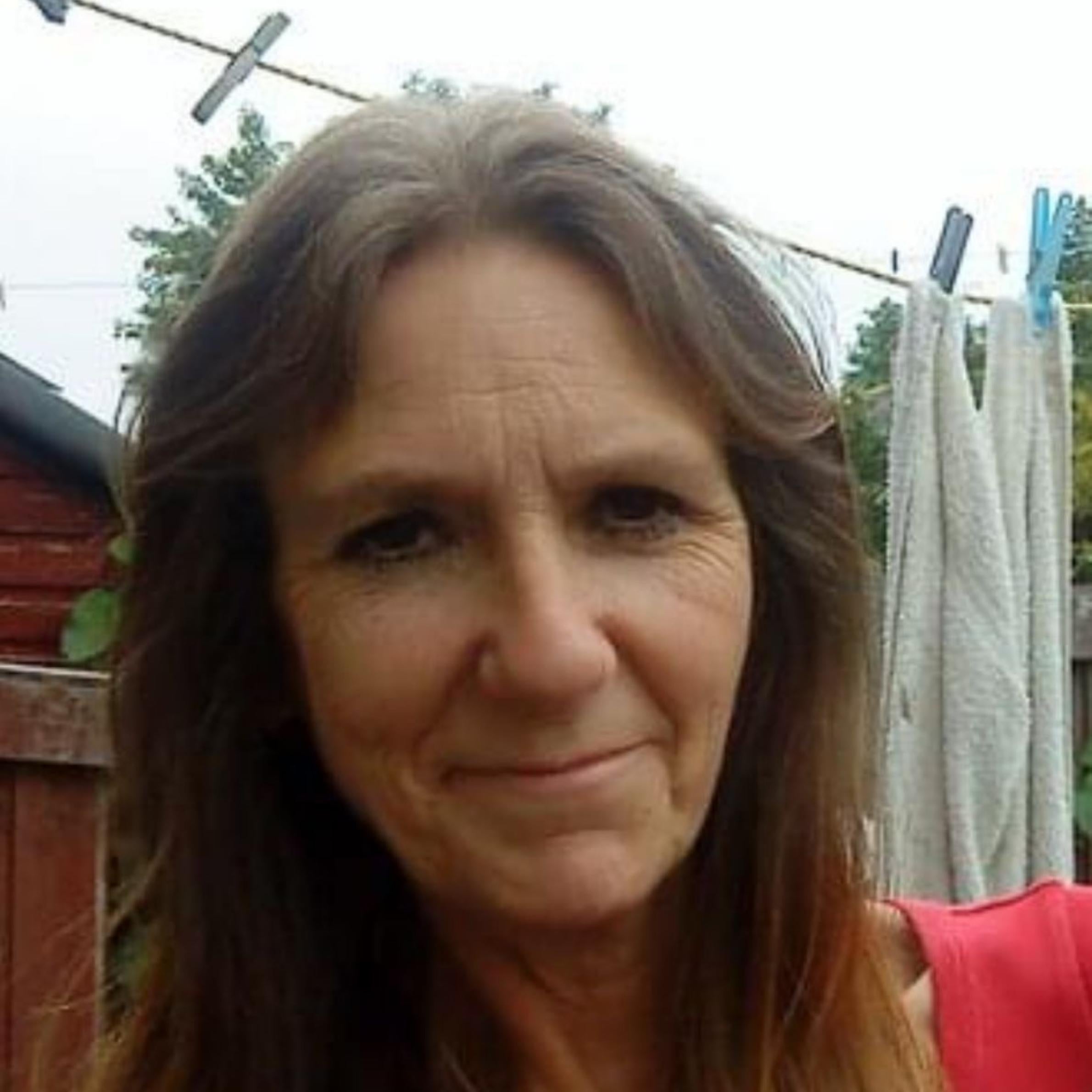 Lady with long brown hair smiling with closed mouth. shes wearing a red top and stood in front of a washing line and brown shed
