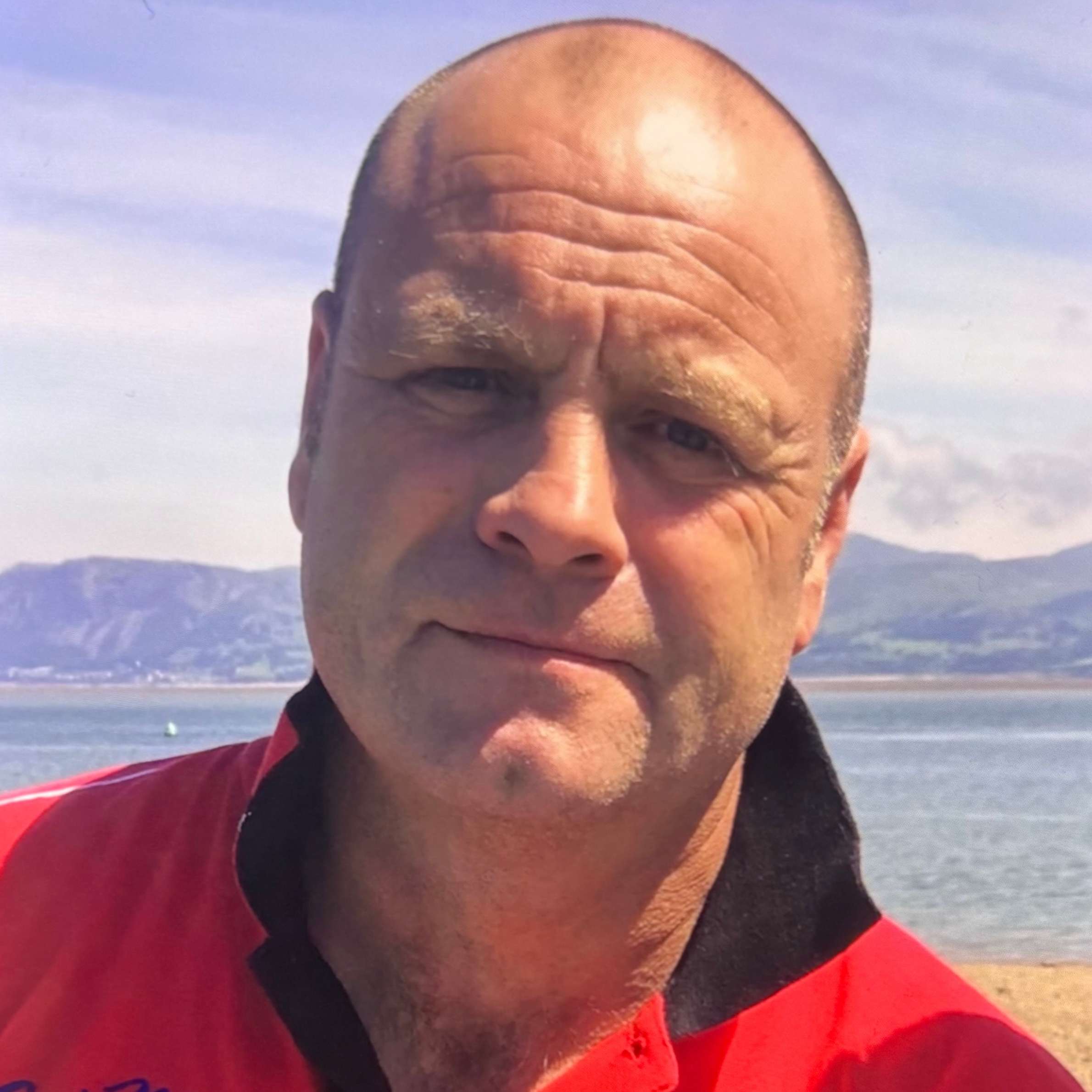 A photo of a man in a red jacket. There is a beach and hills in the background