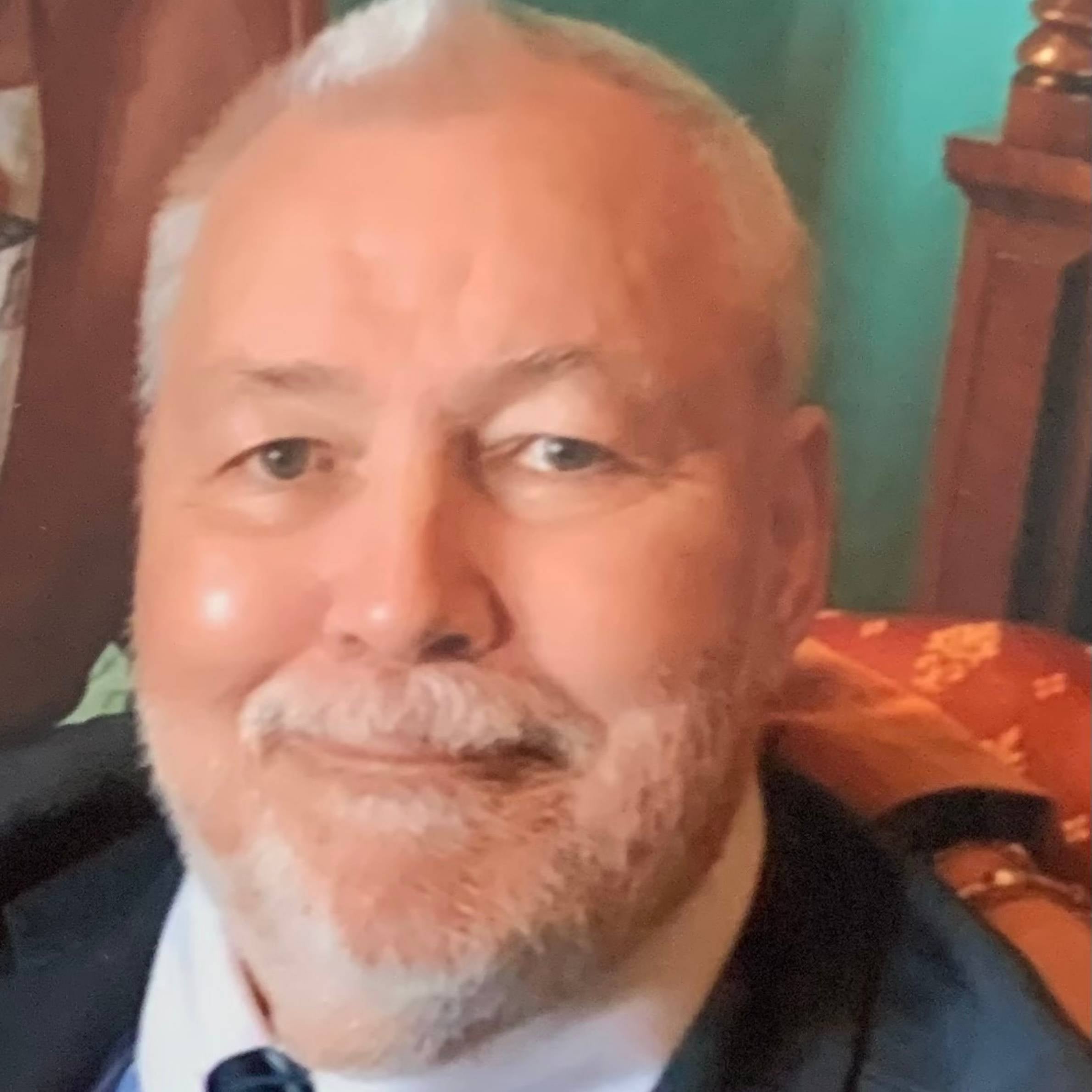 man with white facial hair smiling. he is wearing a collared shirt and tie and blazer. there is a blue wall and wooden furniture behind him