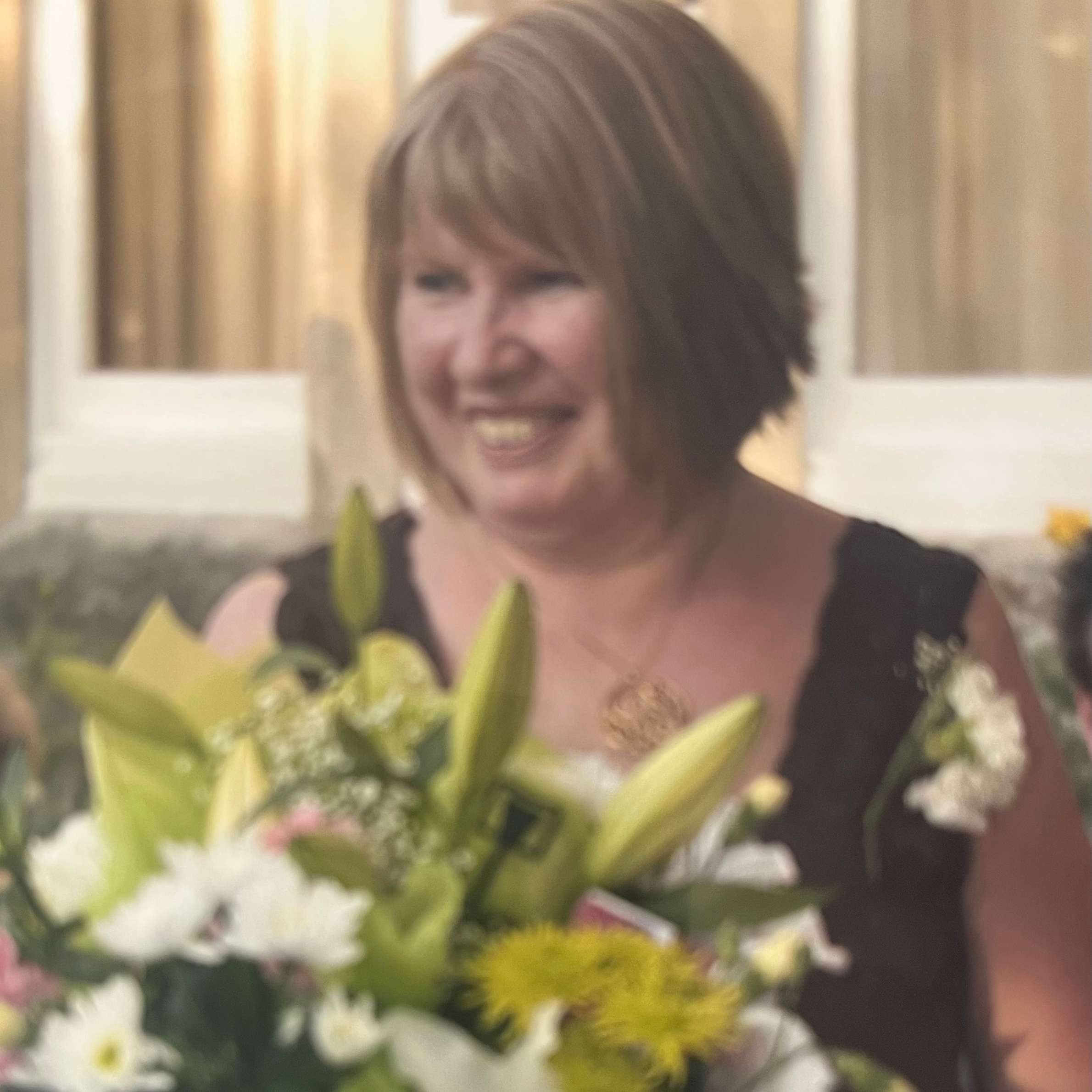 woman with short dark blonde hair smiling standing outside. shes wearing s black top and there is green and white flowers infront of her. behind her there is windows with cream curtains in them