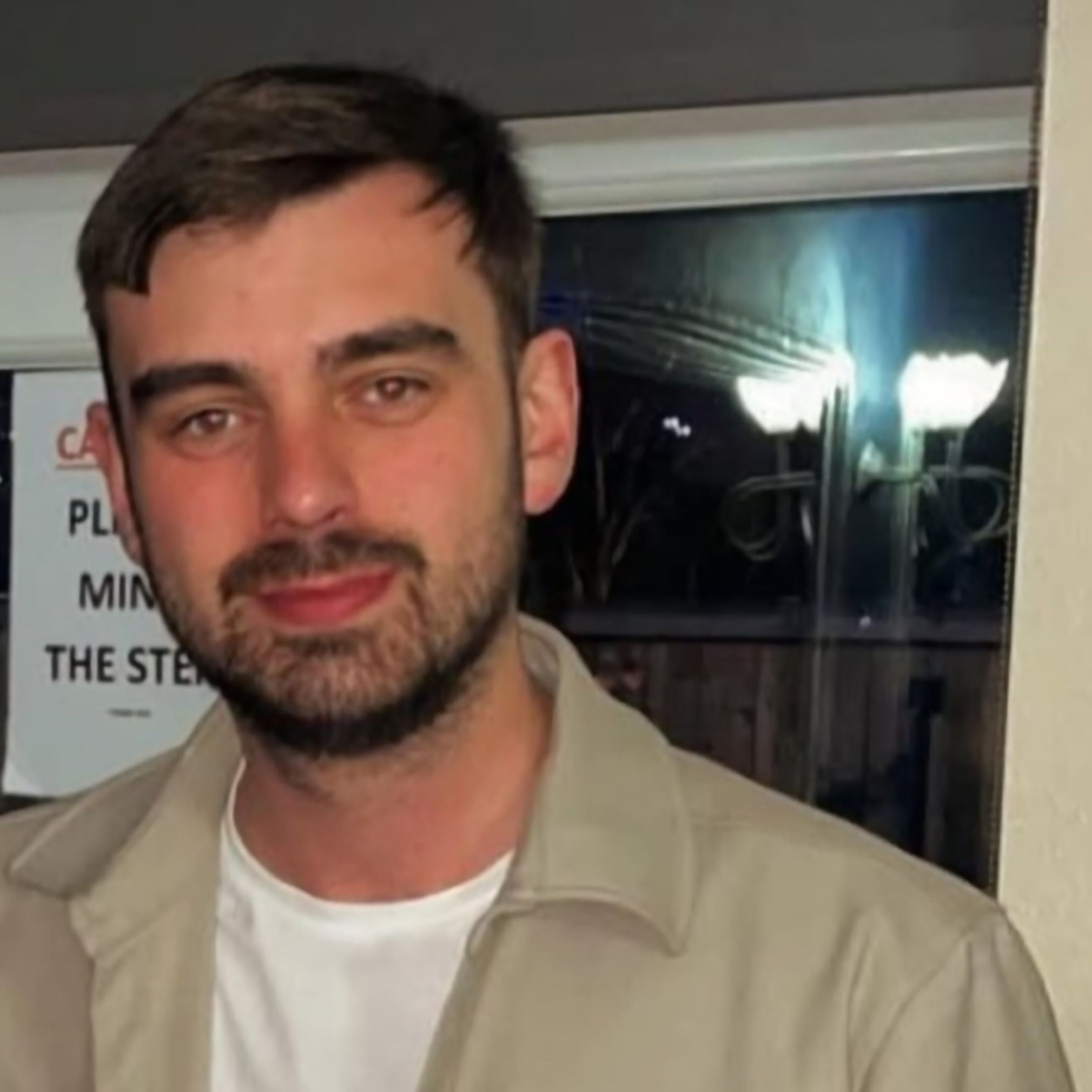 man with black hair and black beard smiling for a photo, he is wearing a white top with a beige shirt on top. there is a window and a white sign in the background