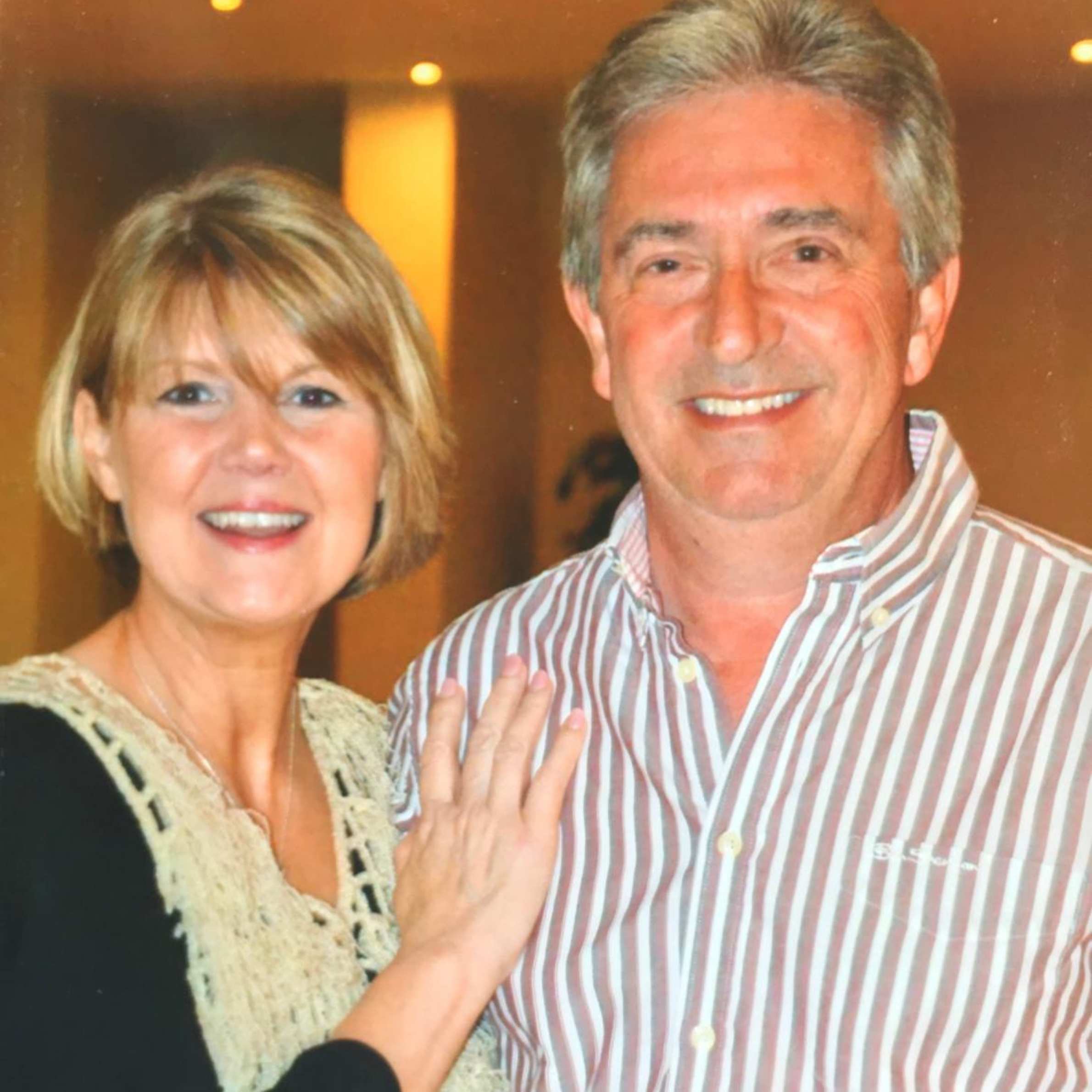 a man and woman smiling for a photo. on the left side there is a woman with short blonde hair wearing a black and white top, she has her hand resting on the mans chest. the man on the right side has grey hair and is wearing a red and white striped shirt