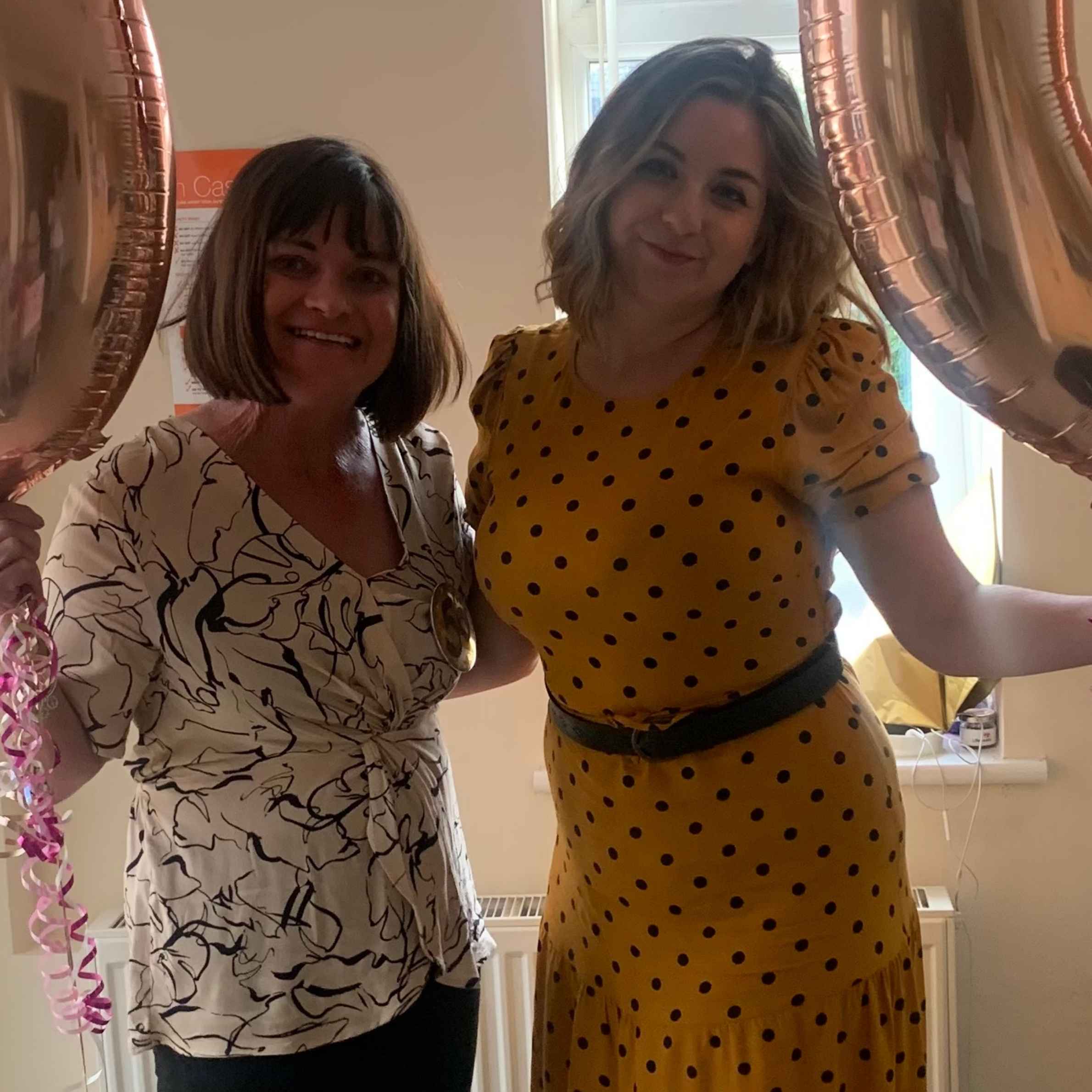 the woman on the left is wearing a white top with a black swirl pattern on it and has short brown hair she is also holding a balloon. the woman on the right is wearing a yellow dress with black dots and has short blonde hair 
